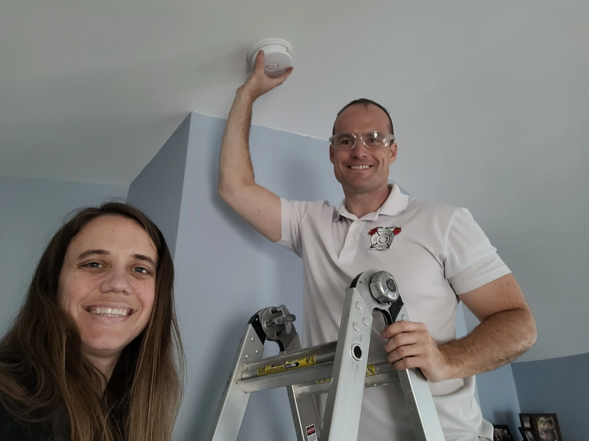 Two people installing a smoke alarm on a ceiling, one on a ladder and one smiling at the camera.