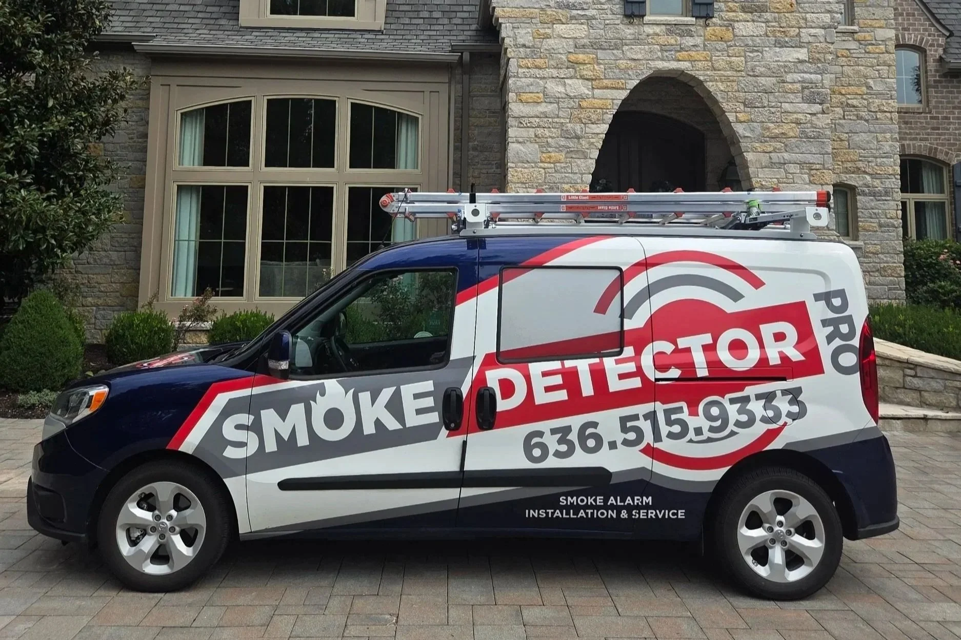 A vehicle parked in front of a house, branded with the logo and contact information of a smoke detection service company. The vehicle has ladders on top and text that reads 'Smoke Detector PRO,' 'Smoke Alarm Installation & Service,' and a phone number.