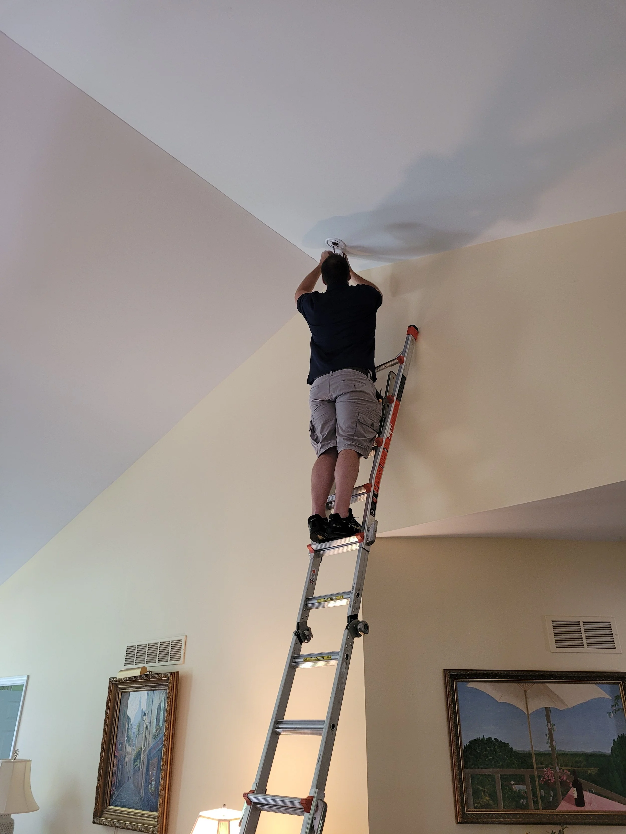 Man standing on a ladder, fixing or installing a ceiling light fixture in a room with beige walls and framed artwork.