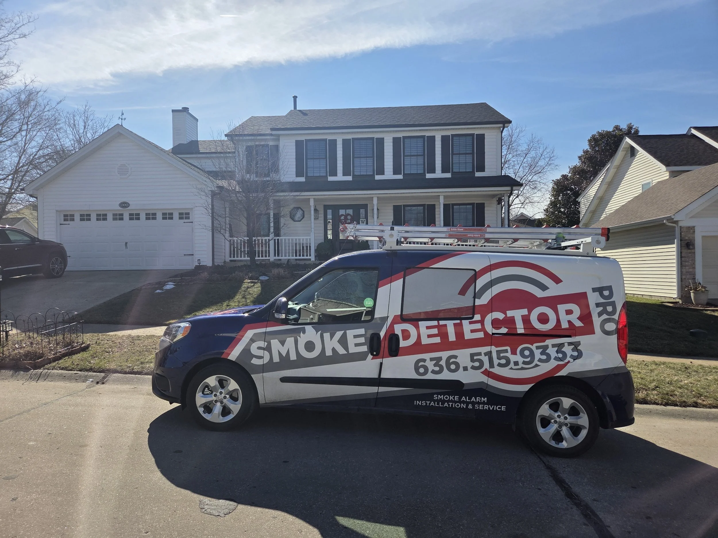 A residential house with a white exterior, dark shutters, and a front porch decorated with holiday wreaths. In the foreground, a service van labeled 'smoke detector' with a ladder on top is parked on the street.