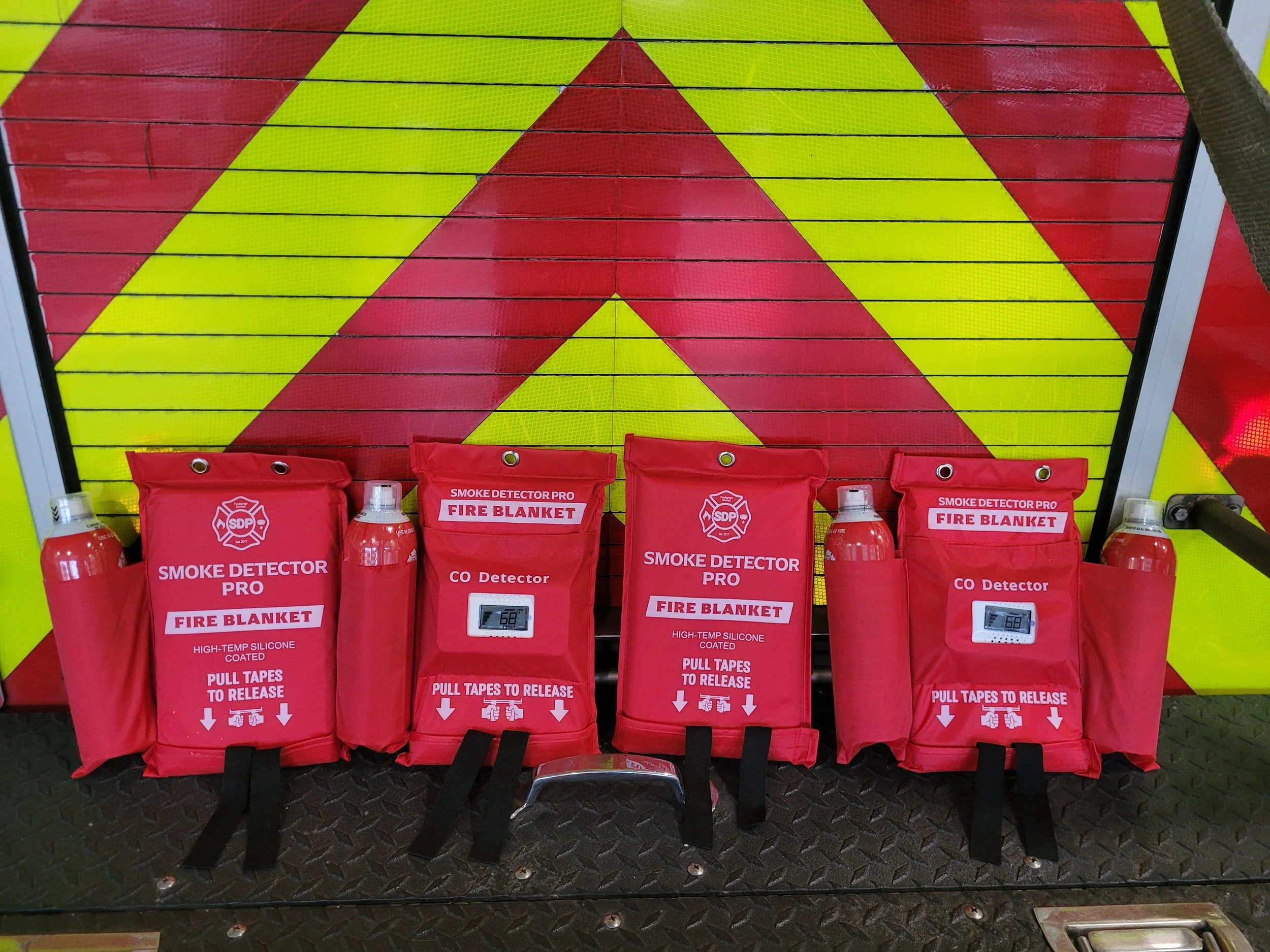 Display of emergency fire safety equipment including red fire blankets, smoke detectors, and fire extinguishers with instructions, set against a yellow and red safety-themed wall.