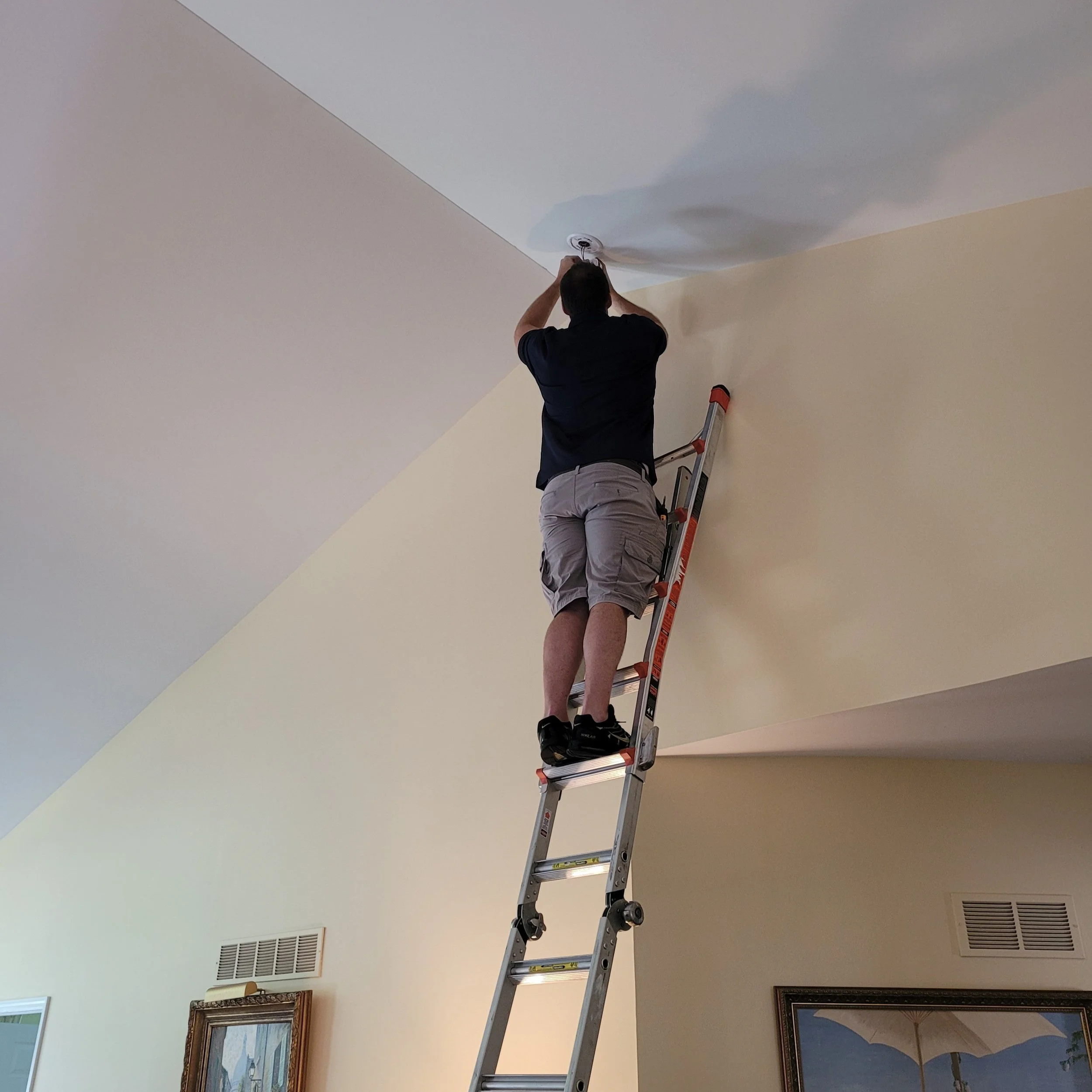 A man on a high ceiling ladder installing a smoke alarm and carbon monoxide alarm.