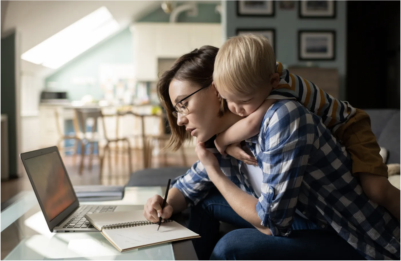 A mother working at a desk while her young child leans on her shoulder