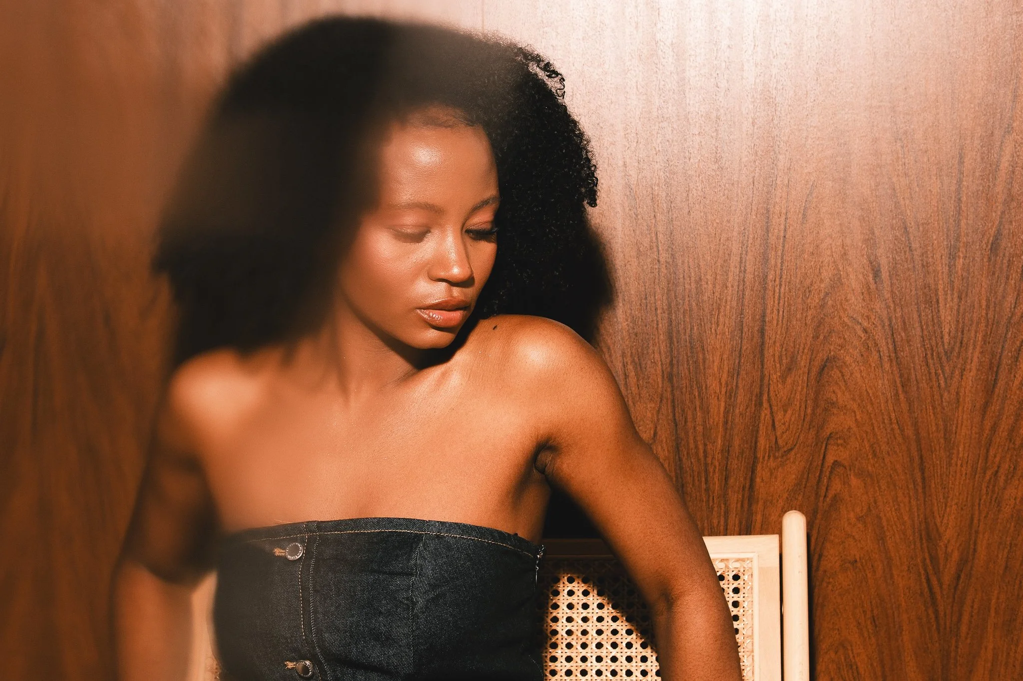 A woman with dark curly hair and smooth skin, sitting against a wooden wall, looking down with her eyes closed, wearing a strapless black top.