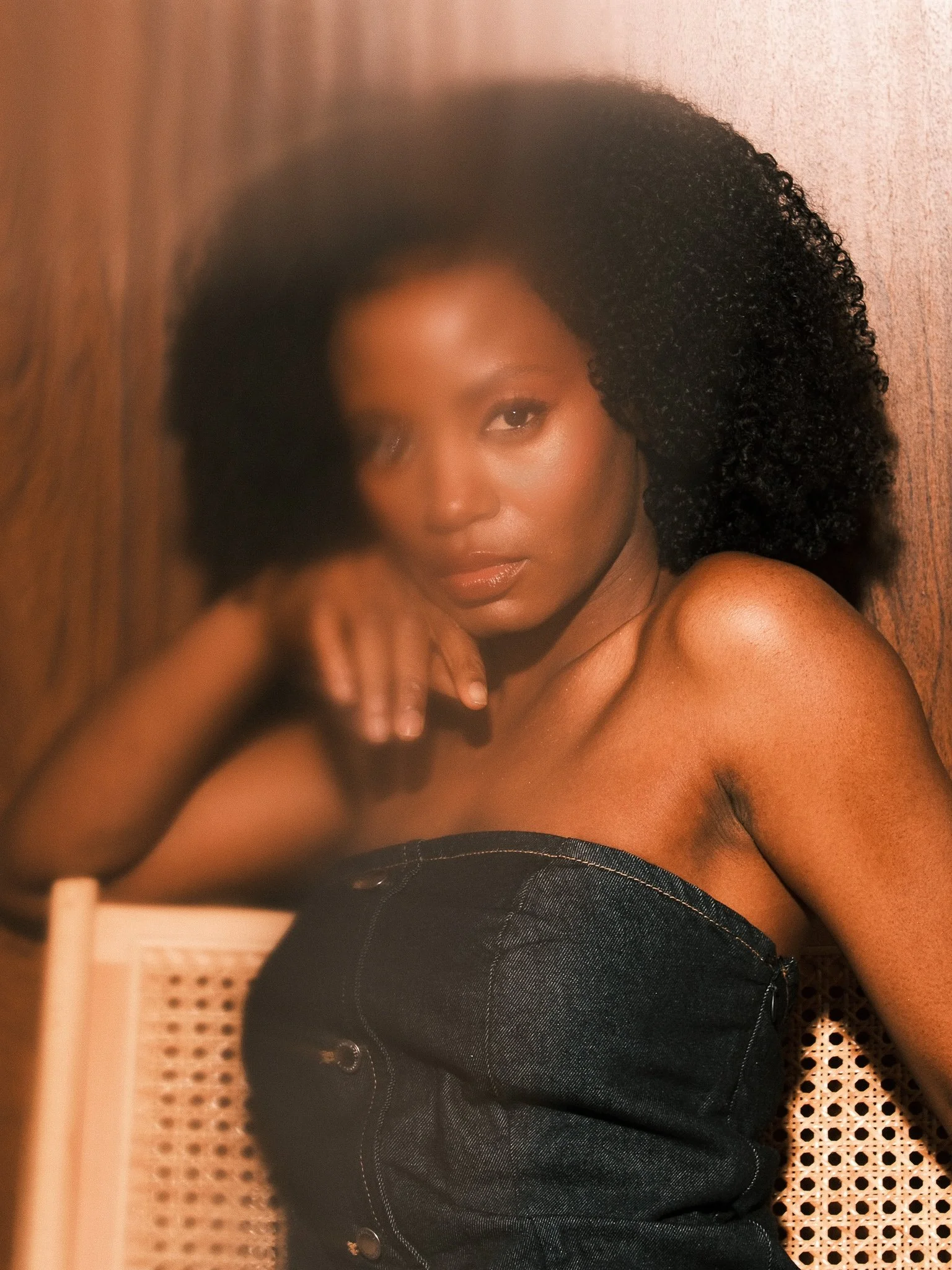 A woman with natural curly hair and smooth skin, sitting against a wooden wall and a woven chair, wearing a black strapless top.