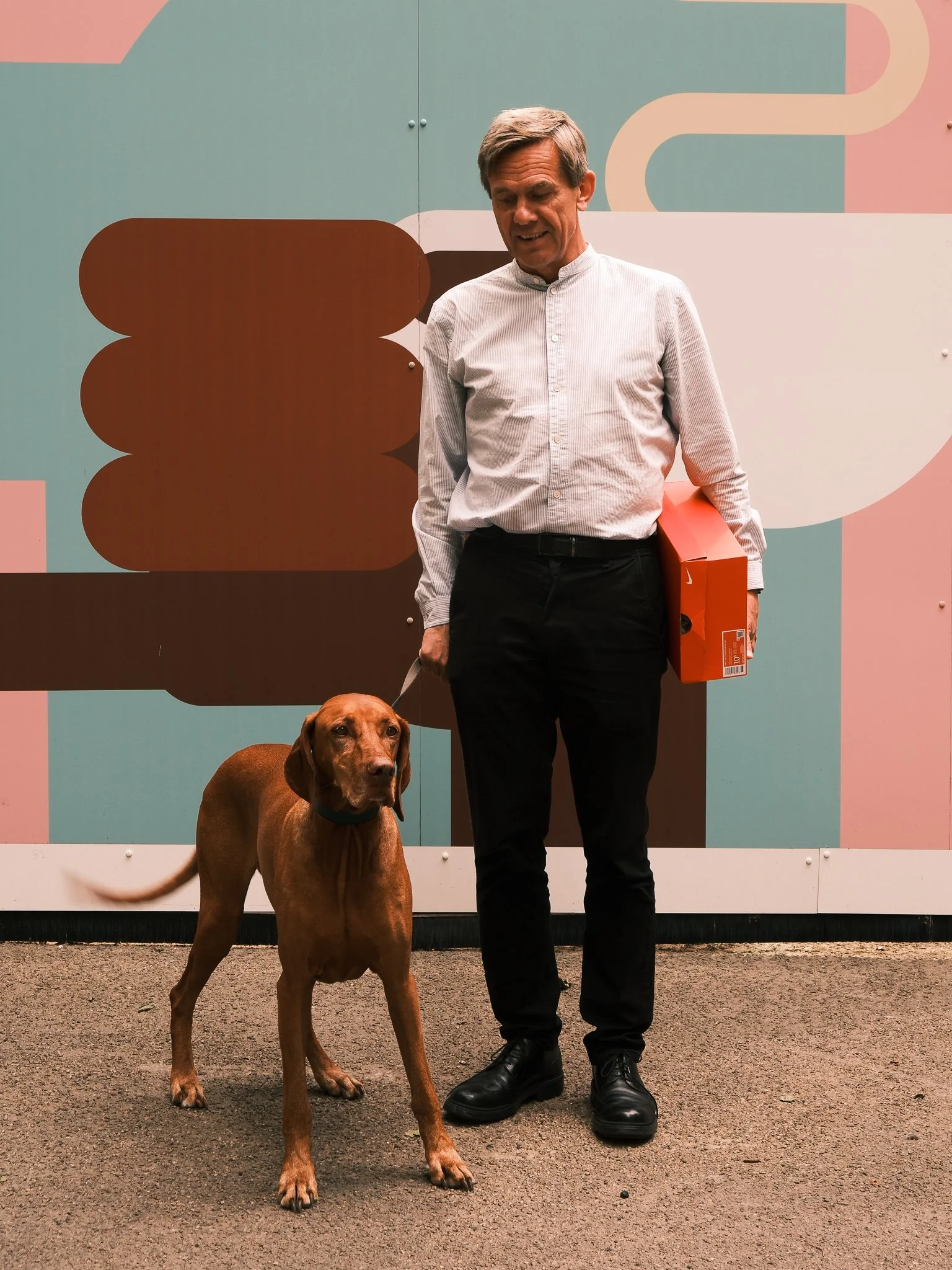 A man in a white dress shirt and black pants holding a red box, standing next to a brown dog on a leash, in front of a colorful geometric background.