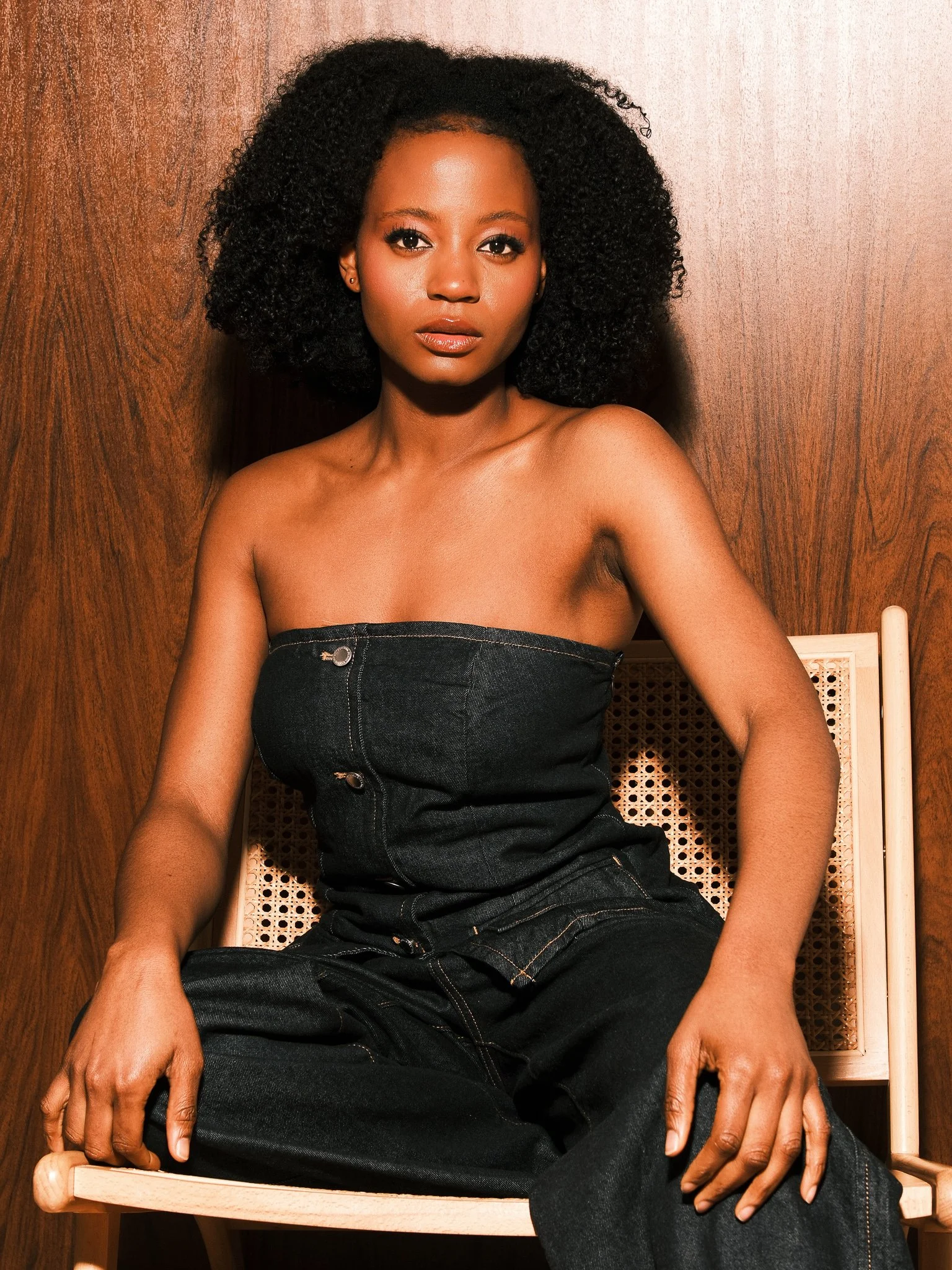 A woman with dark, curly hair and medium brown skin, seated on a wooden chair with a woven cane backrest, against a wooden wall background, wearing a strapless black top and black pants.