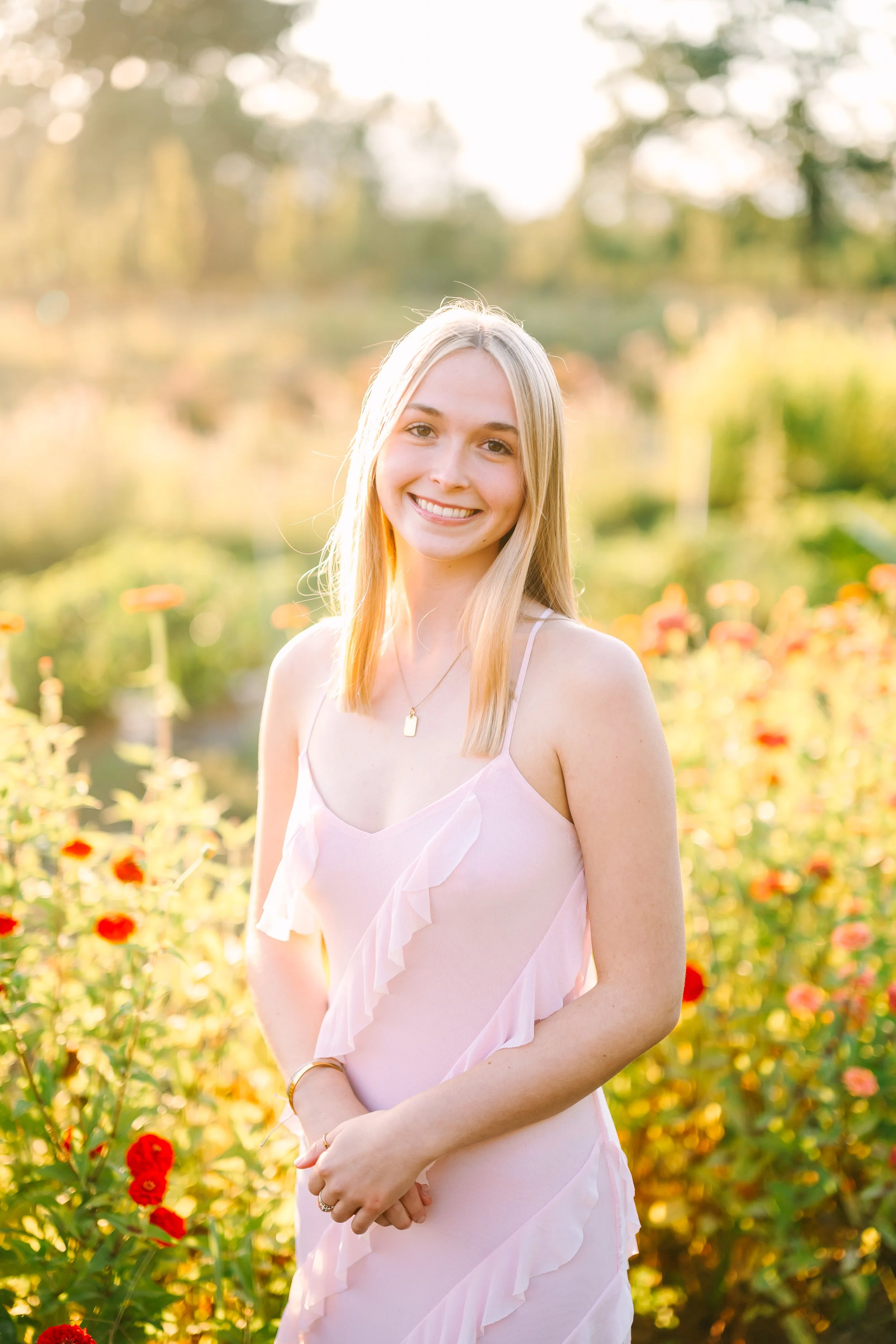 A woman with long, wavy brown hair smiling outdoors in a wooded area with green trees and leaves in the background, wearing a white jacket over a brown shirt and layered necklaces.
