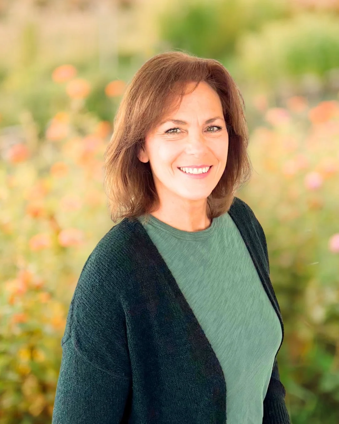 A woman with shoulder-length brown hair smiling outdoors, wearing a green shirt and a black cardigan, with a blurred natural background of greenery and flowers.