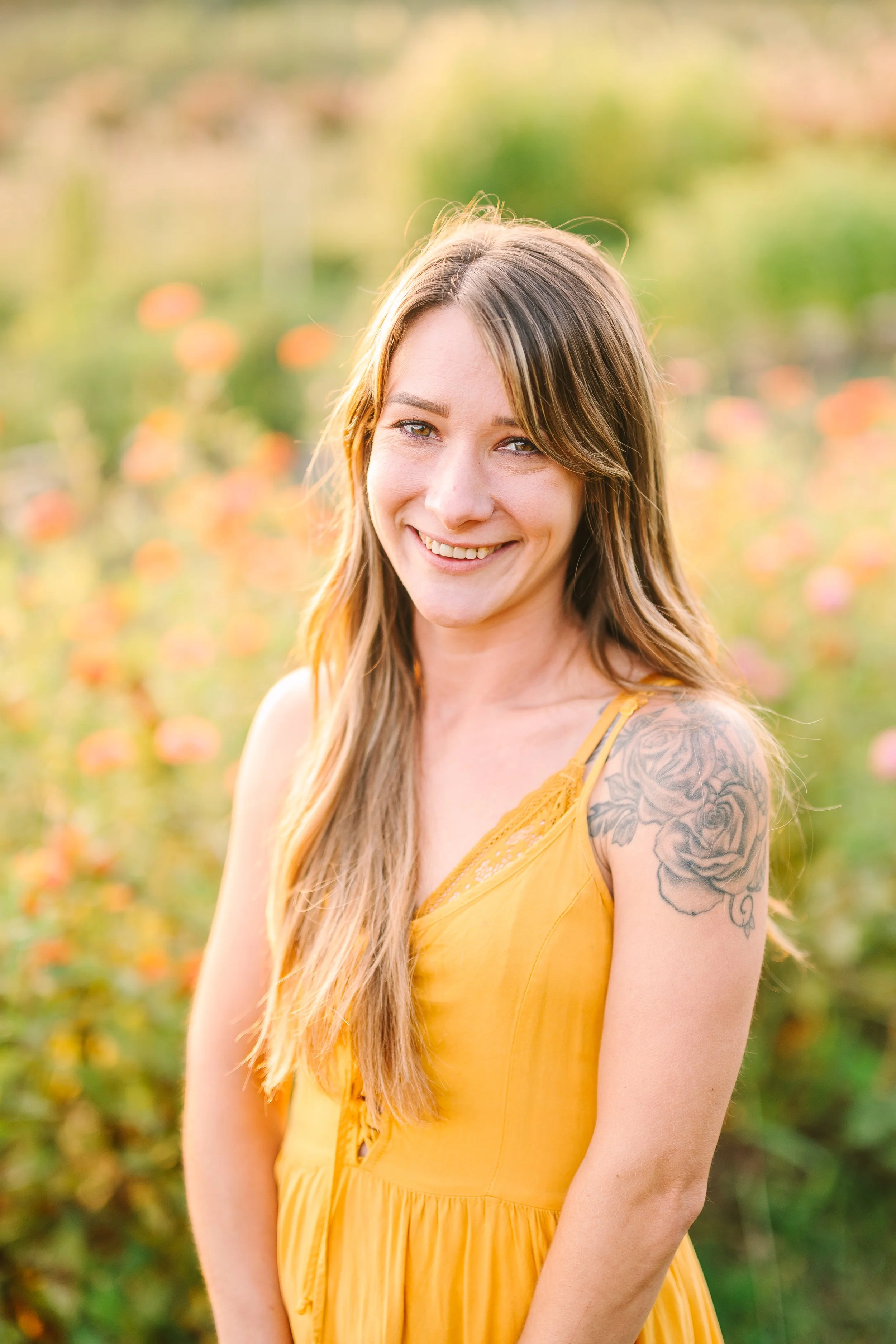 A woman with long light brown hair and a tattoo of roses on her shoulder, smiling while standing in a field of orange and pink flowers.