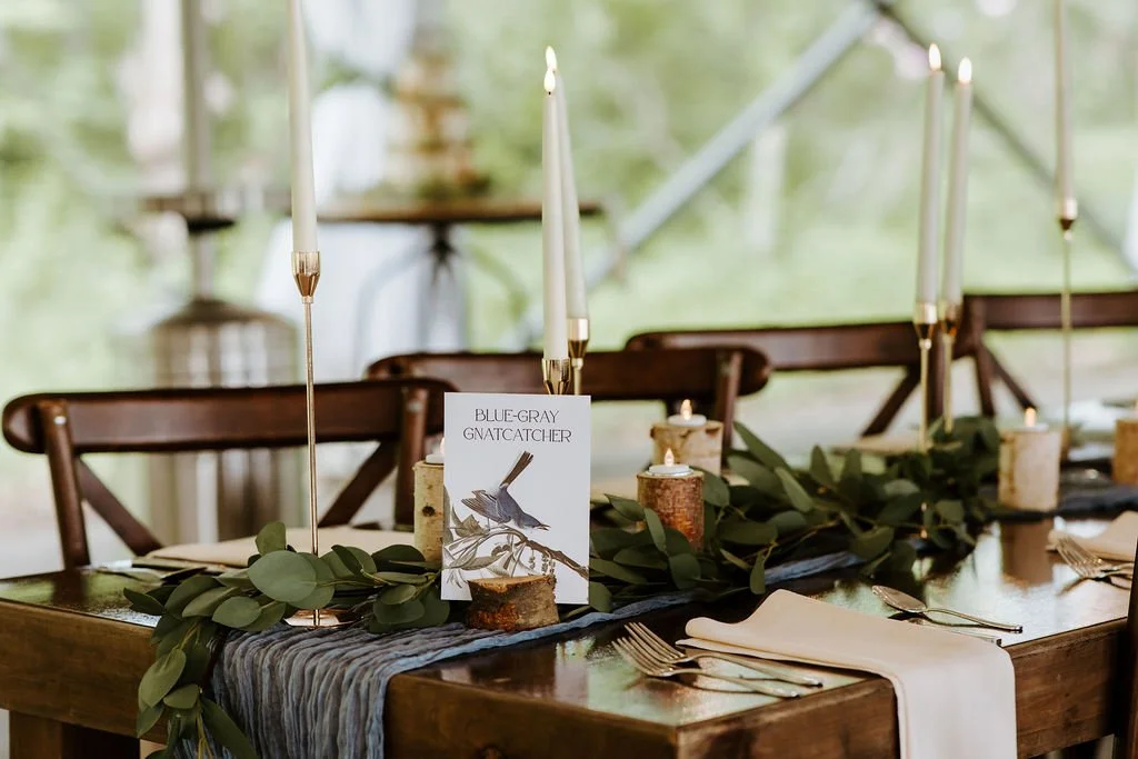 Elegant dining table with greenery, candles, and tall slim candle holders, set with folded white napkins and silverware, in a well-lit room with windows showing trees outside.