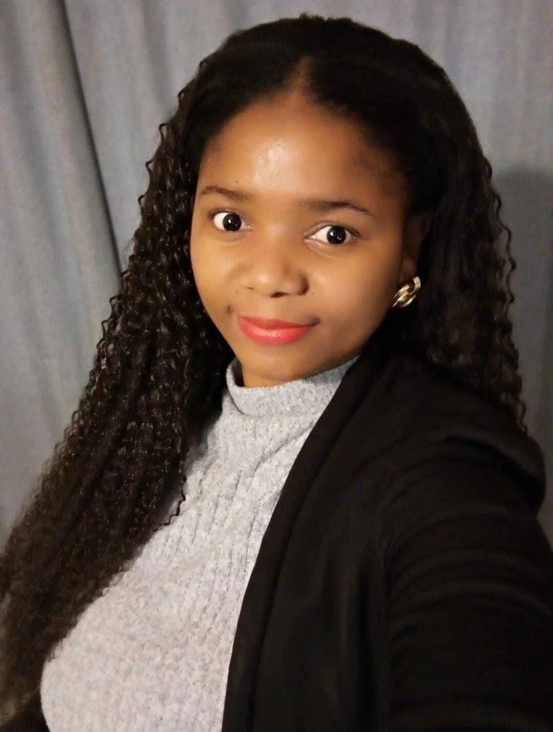 A woman with long, curly dark hair, wearing a gray turtleneck sweater, a black blazer, and gold earrings, taking a selfie in front of a gray curtain.