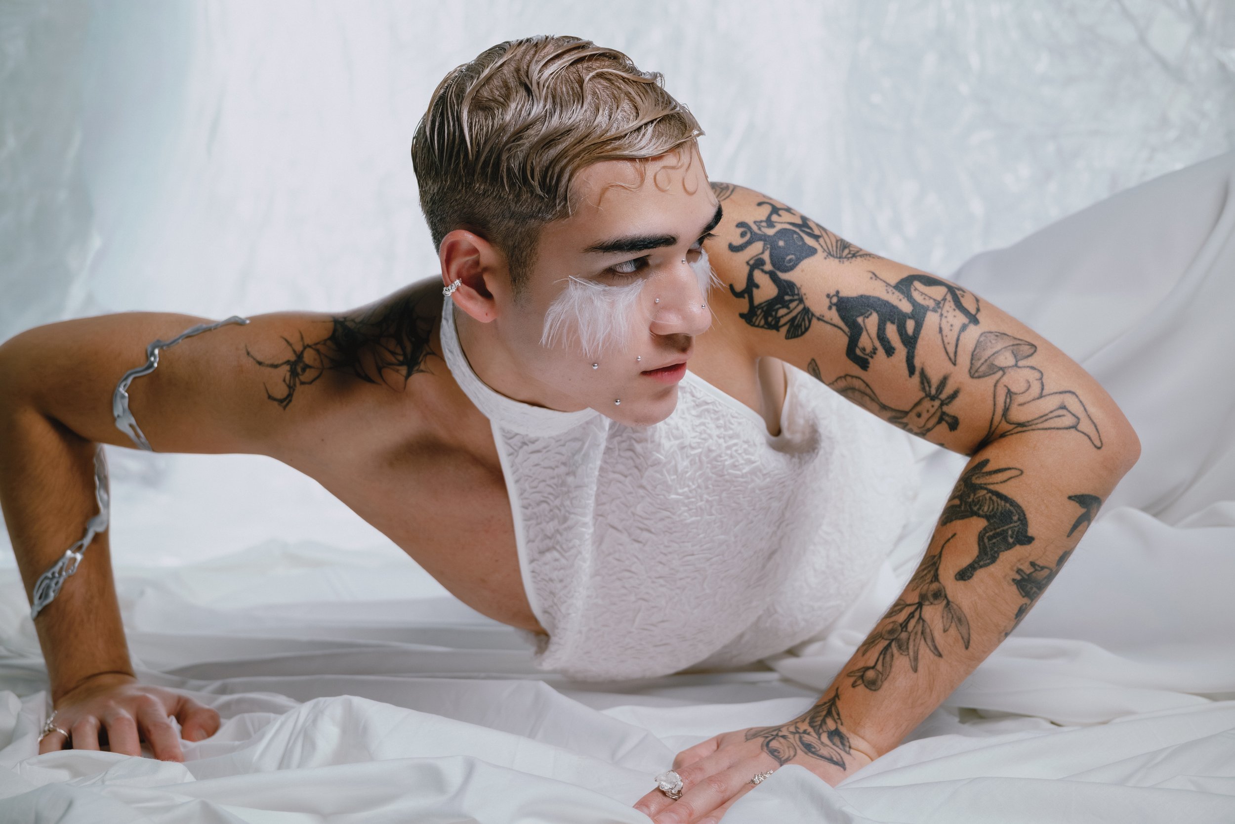 Person with short blonde hair and tattoos lying on white bed, wearing a white textured outfit, with facial piercings and makeup, in a close-up profile shot.