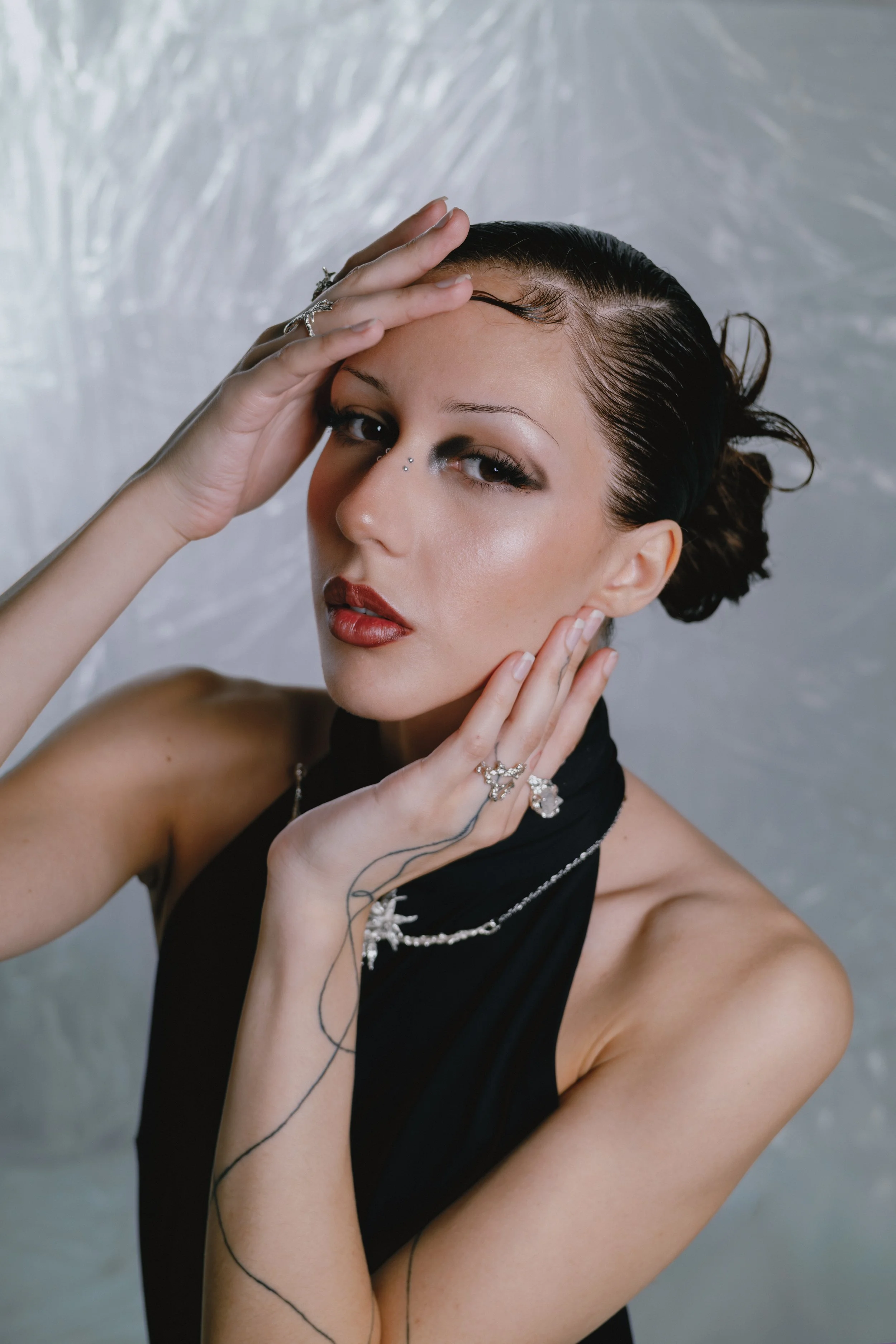 A woman with dark hair styled back, wearing makeup with bold red lipstick, and accessorized with rings and necklaces. She is holding her hand to her face and looking at the camera, with a silver, textured background.