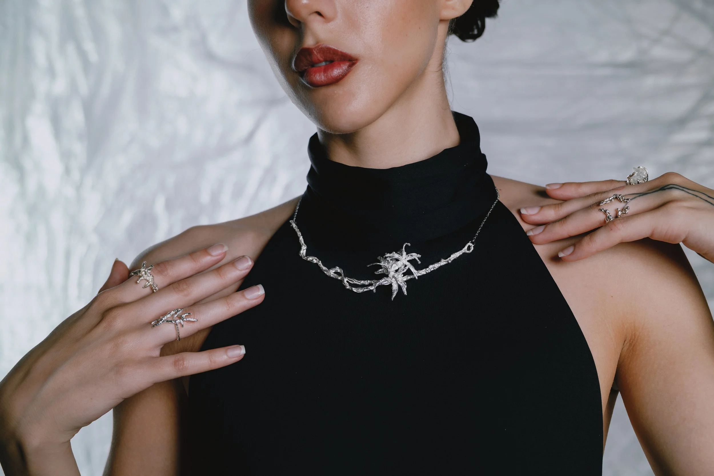 A woman with red lipstick wearing a black turtleneck and silver jewelry, including a necklace and rings, against a light, textured background.