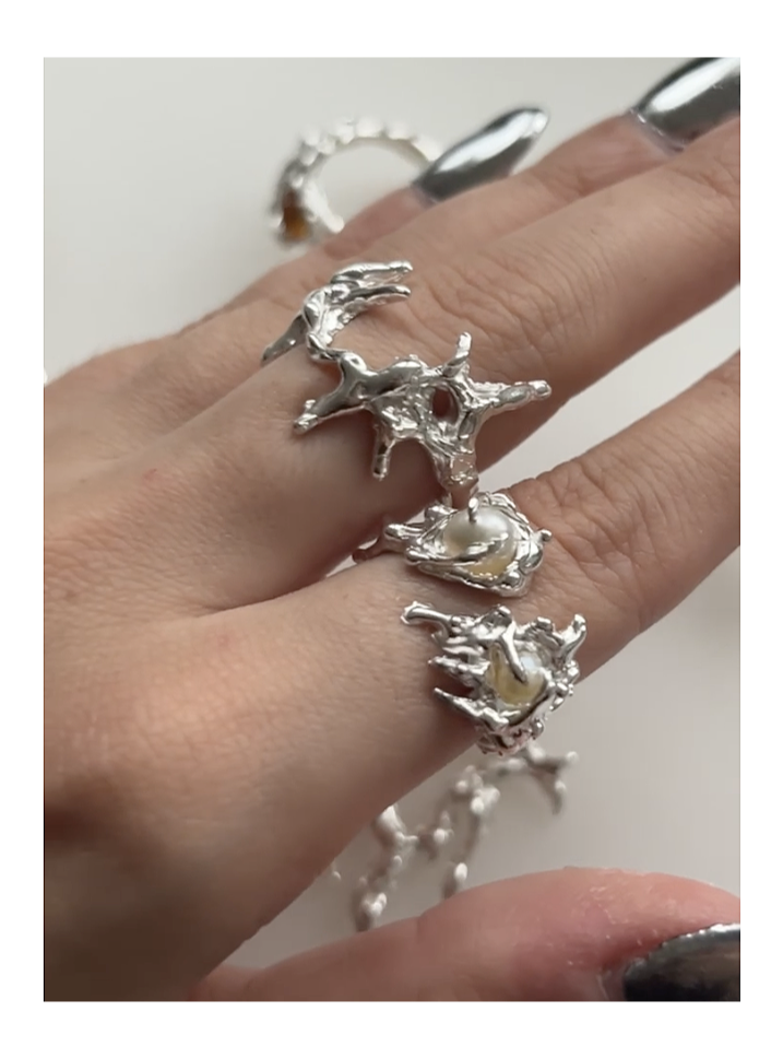 Multiple silver rings with organic, coral-like designs on a person's fingers, some featuring pearls, with a silver chain in the background.