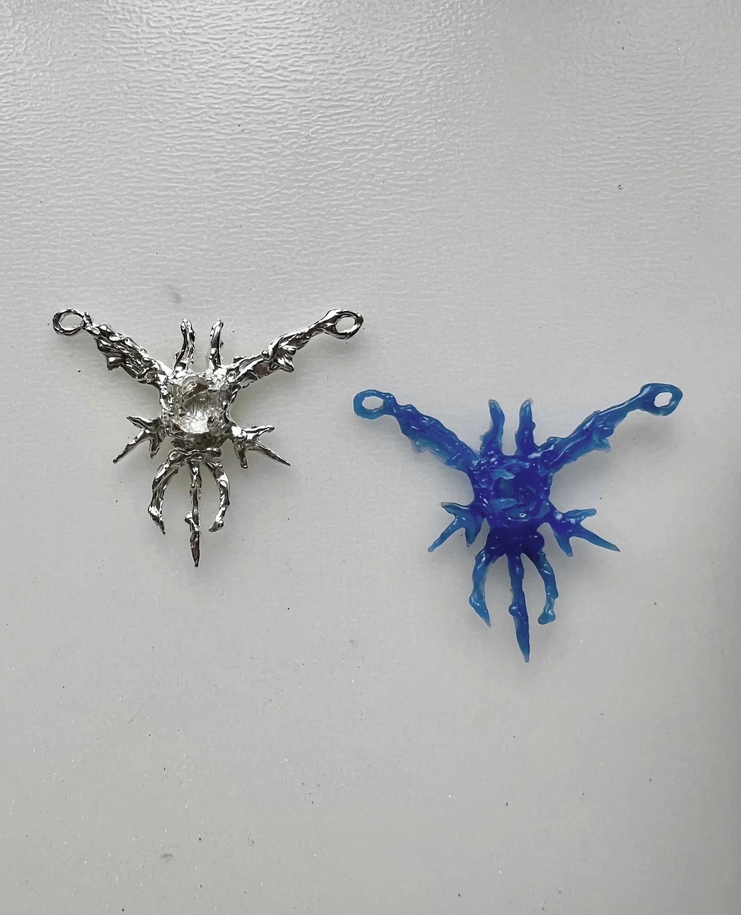 Two jump rings with spider-shaped charms, one made of silver metal and the other of blue glass or plastic, on a white surface.