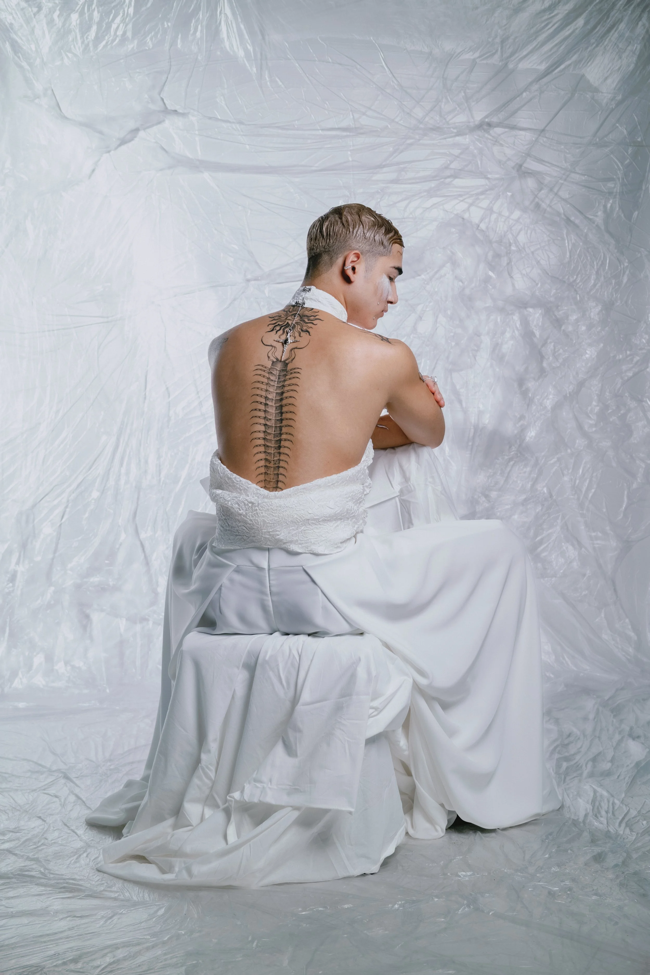 A person with a short haircut, sitting on a white cloth-covered stool, with their back to the camera. They have a large, detailed tattoo of a spine on their back.