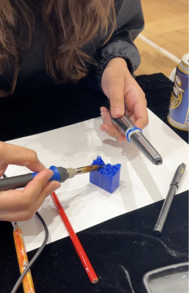 Person holding a soldering iron and soldering a blue 3D-printed object on a white work surface indoors.