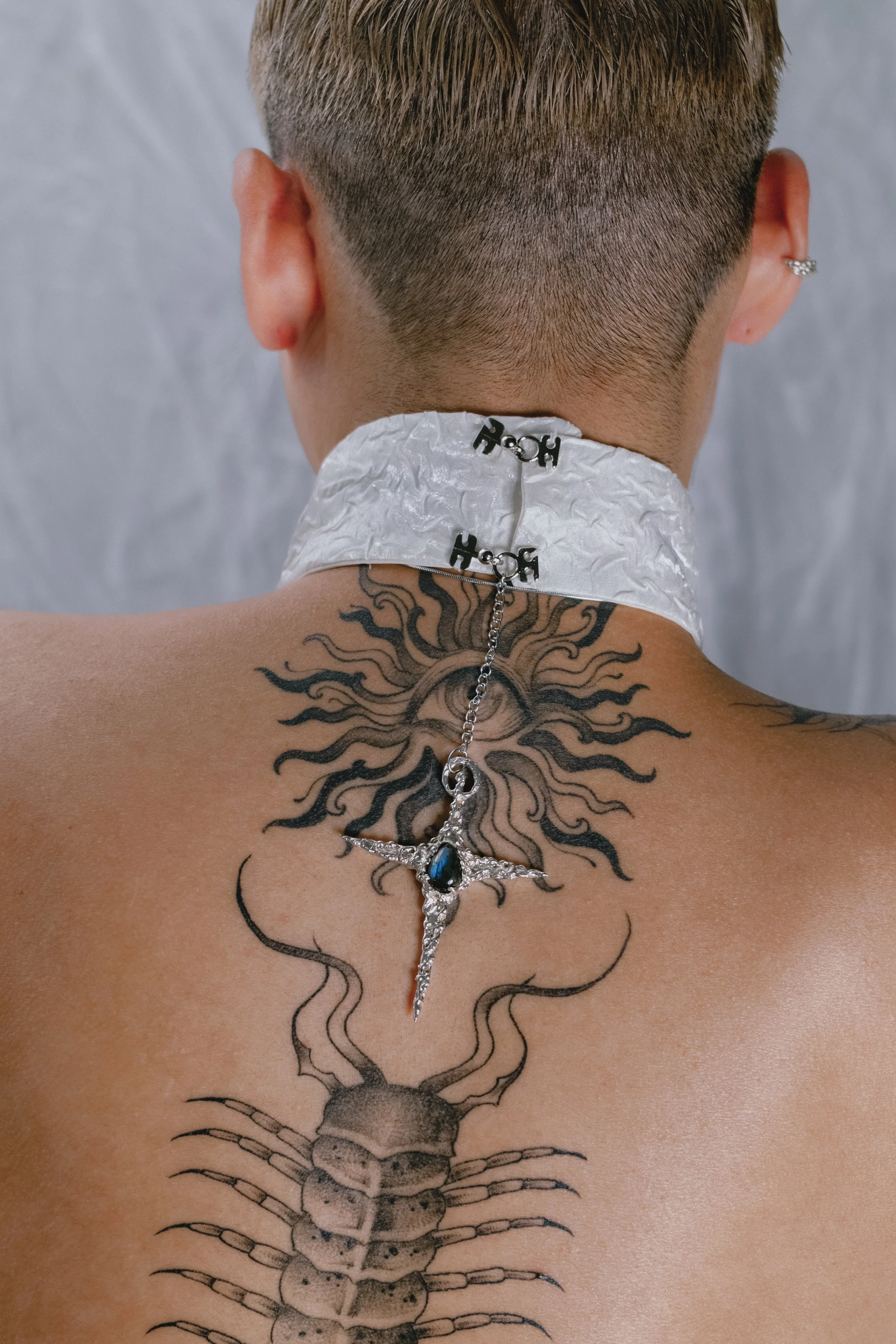 Back of person with short hair, tattoo of a scorpion and a sun with rays, wearing a white choker with black 'ROH' pins and a silver cross necklace with a dark gemstone.
