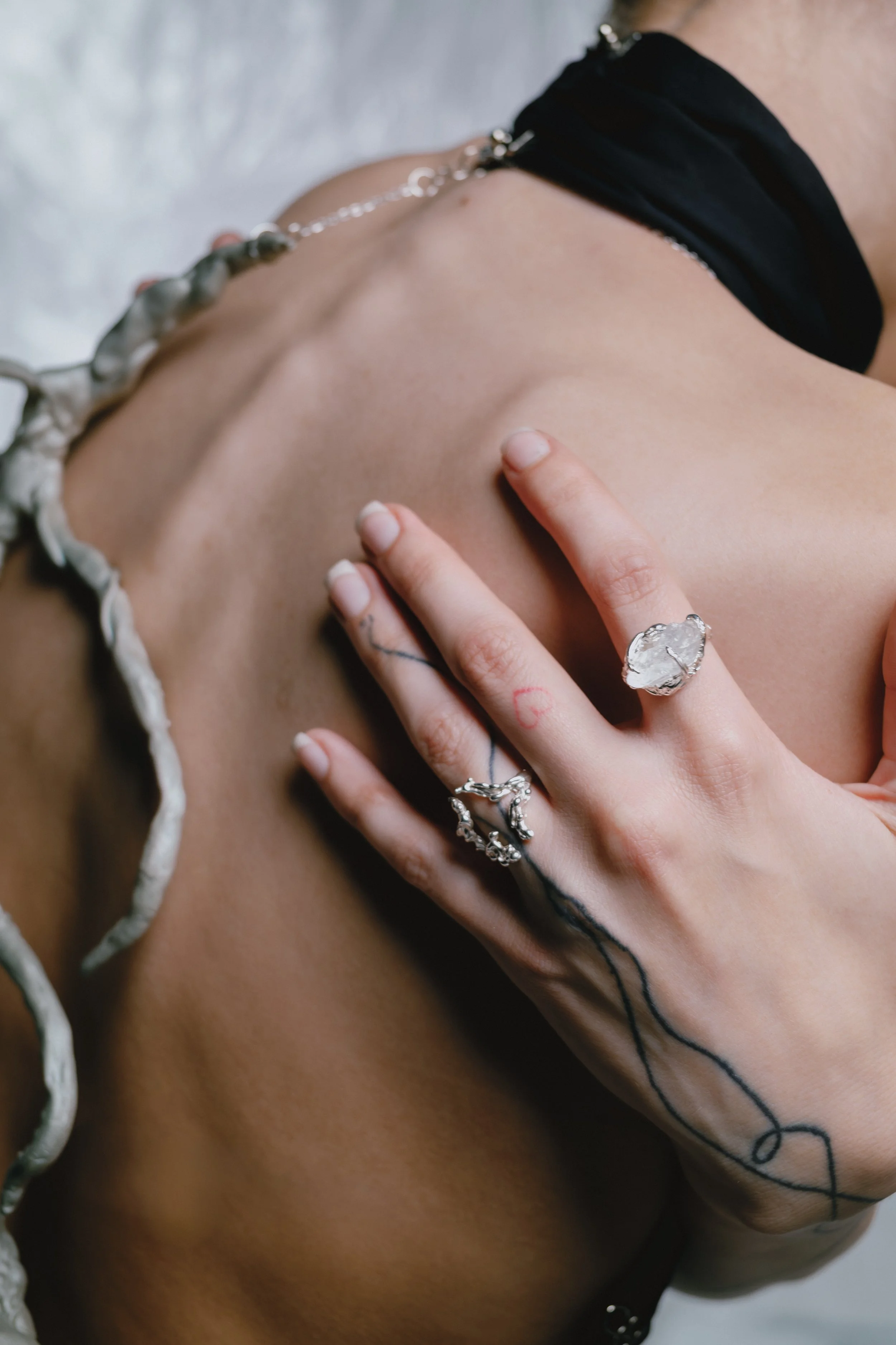 Close-up of a person's hand with rings, resting on their clavicle, showcasing tattoos, with a portion of black clothing and jewelry visible, against a neutral background.