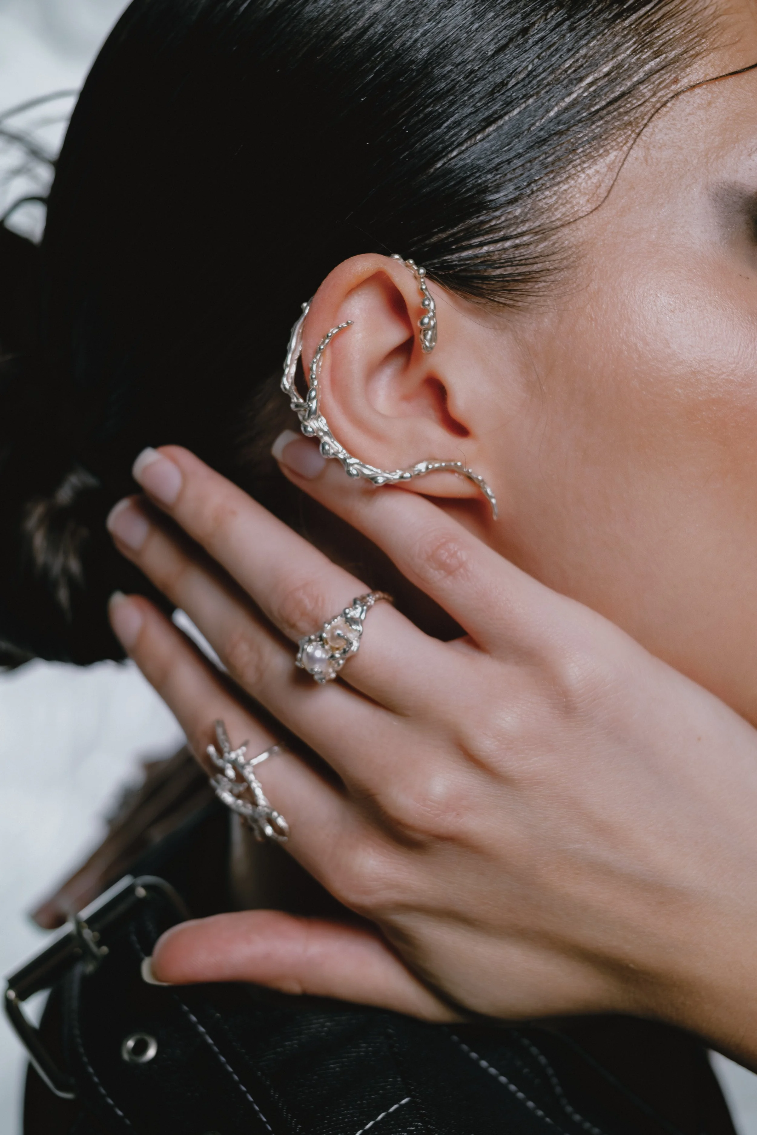 Close-up of a woman with dark hair wearing multiple silver jewelry pieces, including earrings, rings, and ear cuffs, touching her face with her fingers.