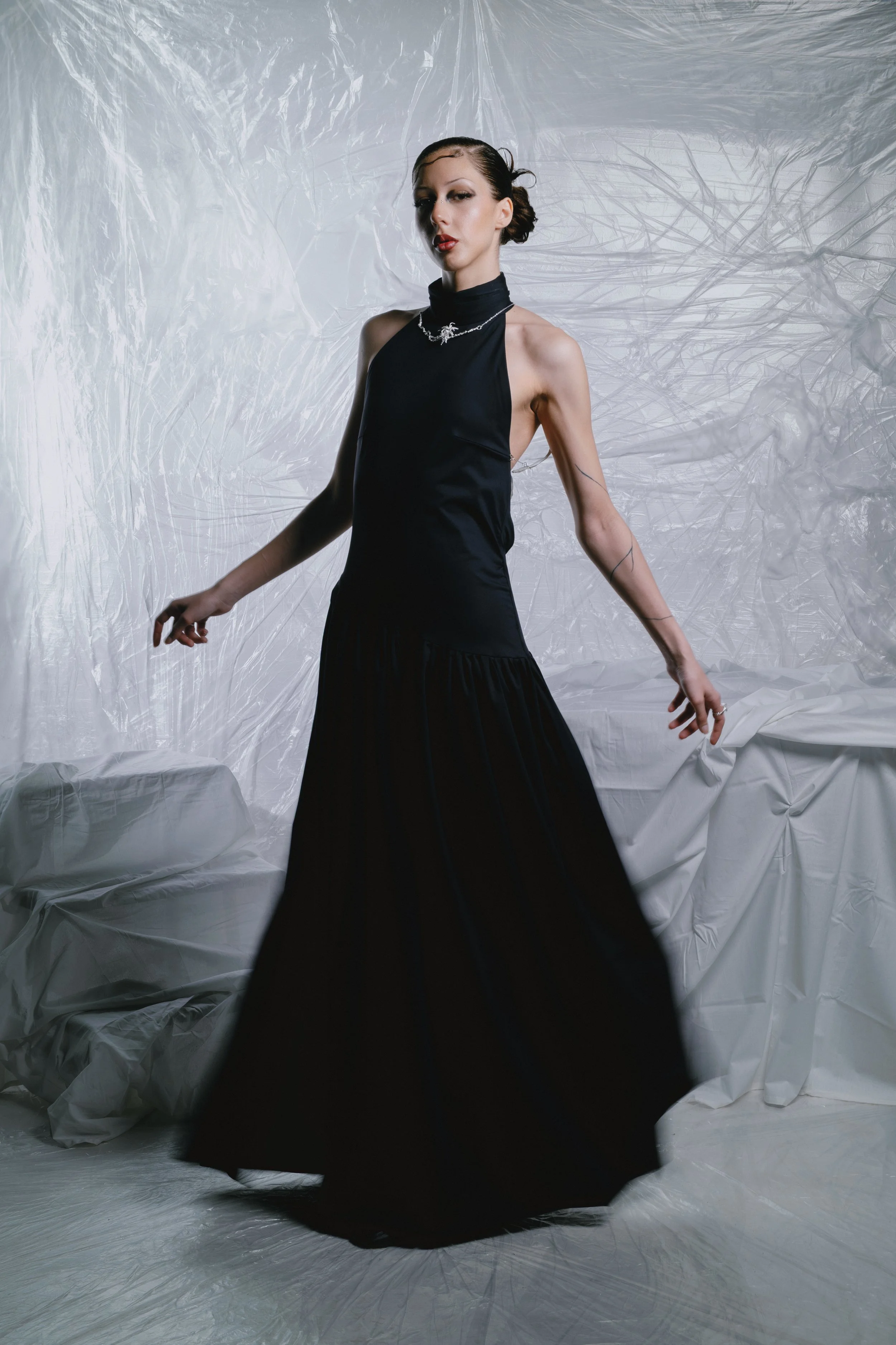 A woman in a long black dress standing in front of a crinkled white background, posing with one arm extended outward and the other hanging down.