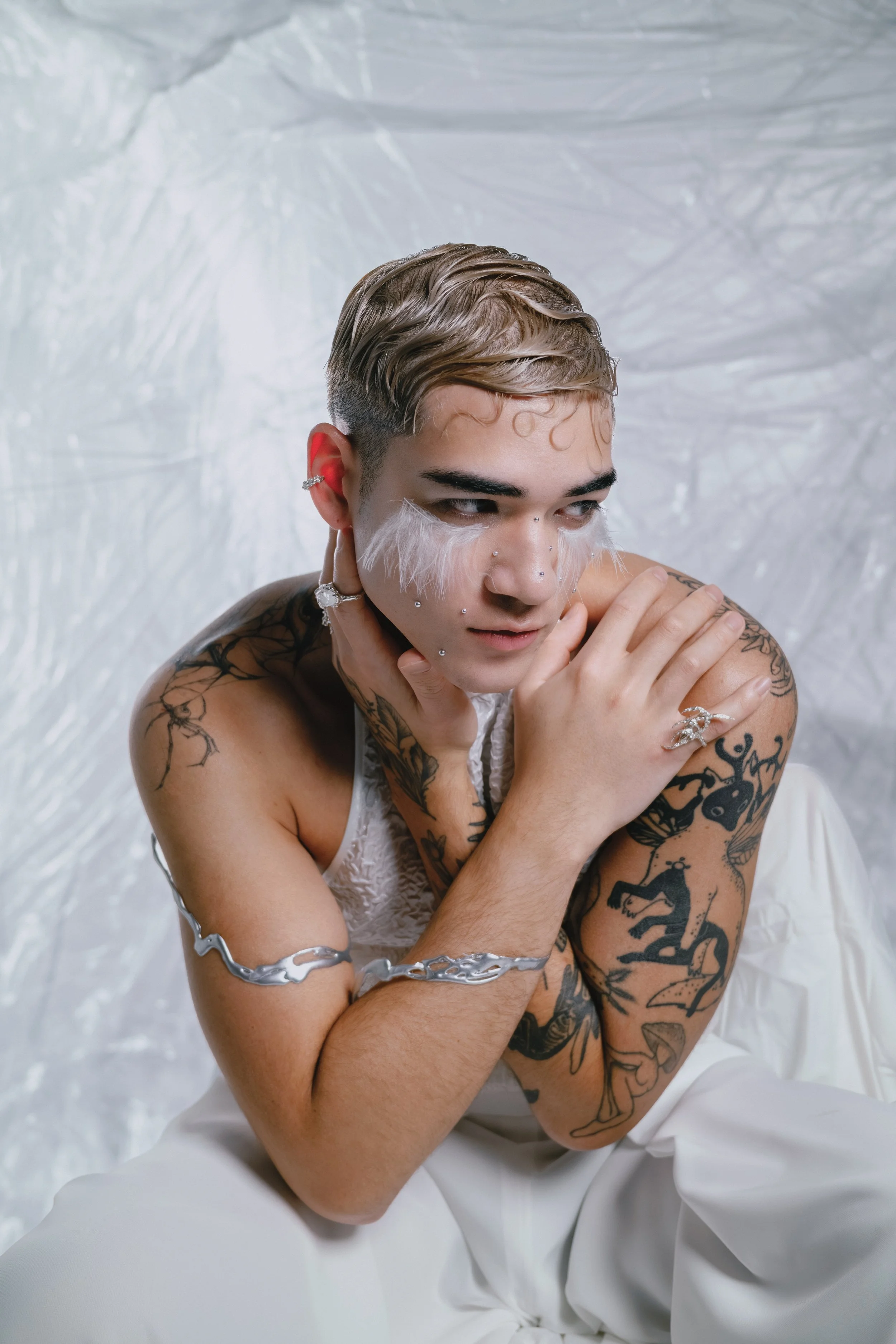 Person with short, slicked-back hair and multiple tattoos, wearing white feather makeup, sitting against a textured white background.