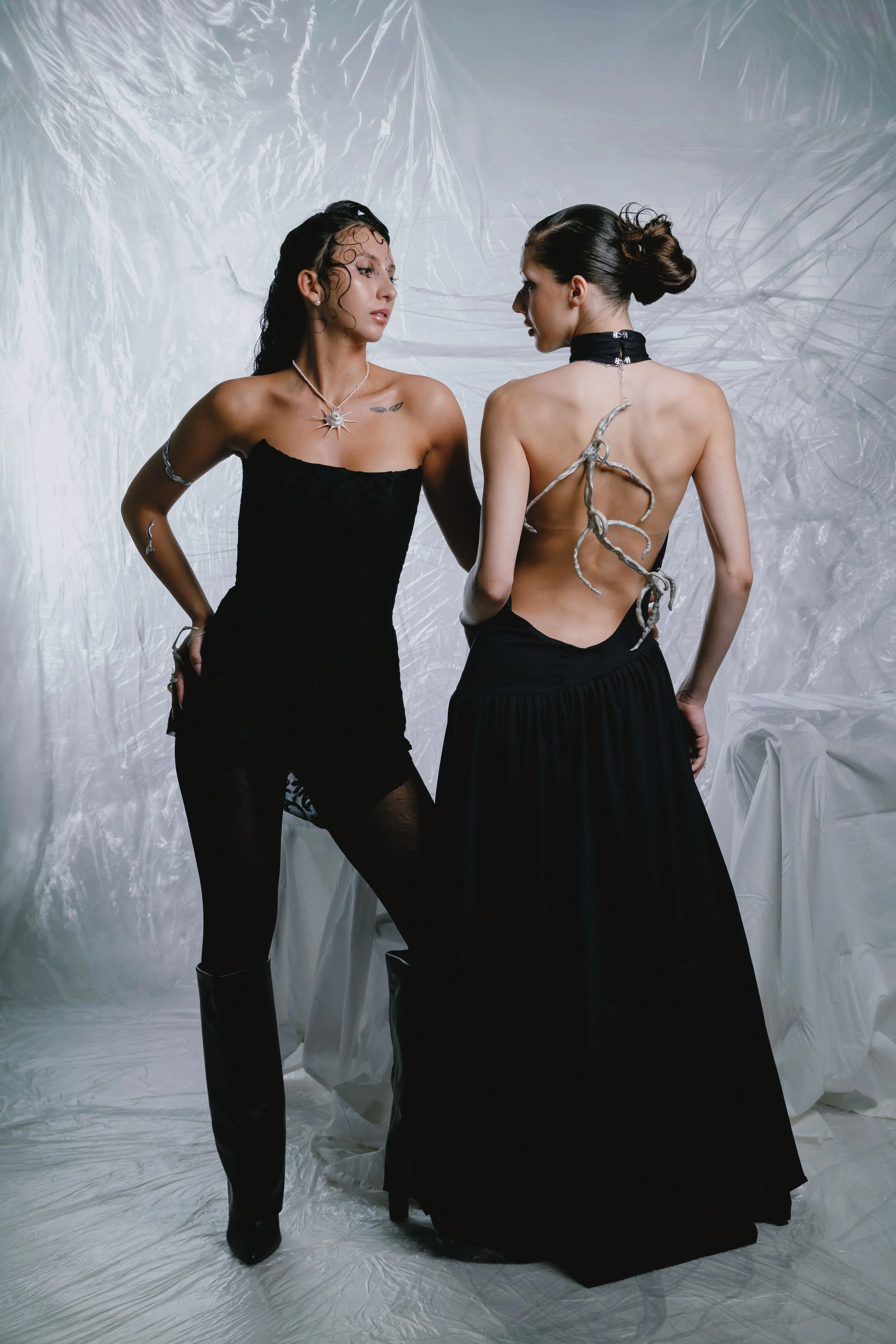 Two women in black dresses posing against a textured white backdrop; one wears a strapless dress, the other a backless gown with a silver snake-like accessory and choker.