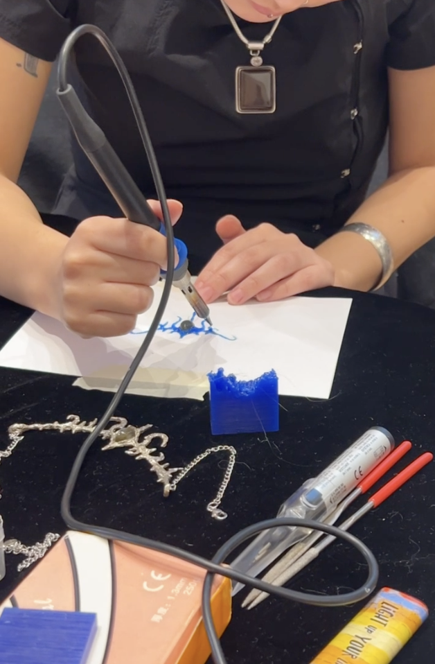 Person creating jewelry using a soldering iron, with jewelry pieces and tools on the table.