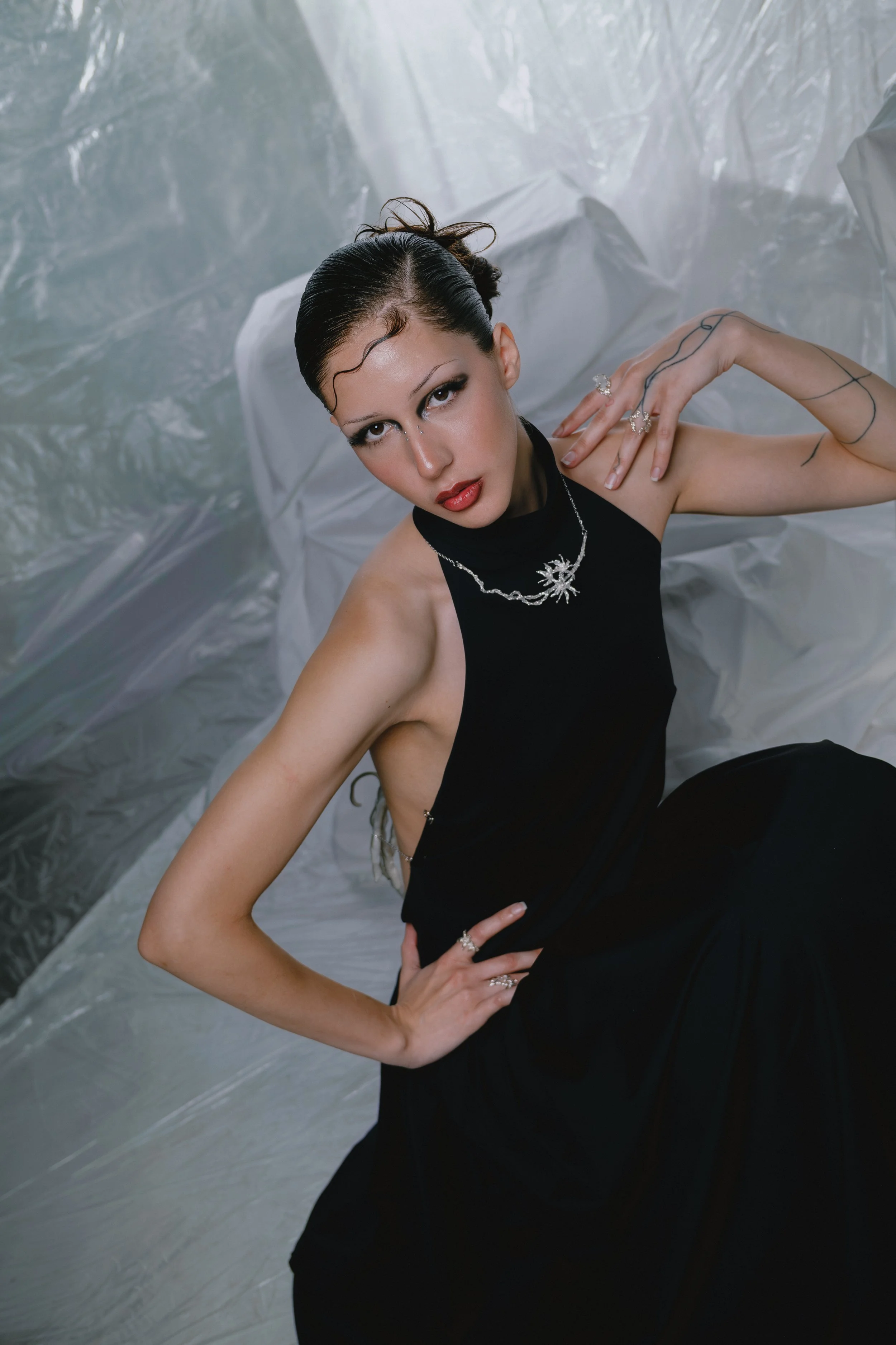 A woman in a black dress sitting on a silver surface with a plastic backdrop, looking up at the camera, wearing jewelry including a necklace, rings, and earrings.
