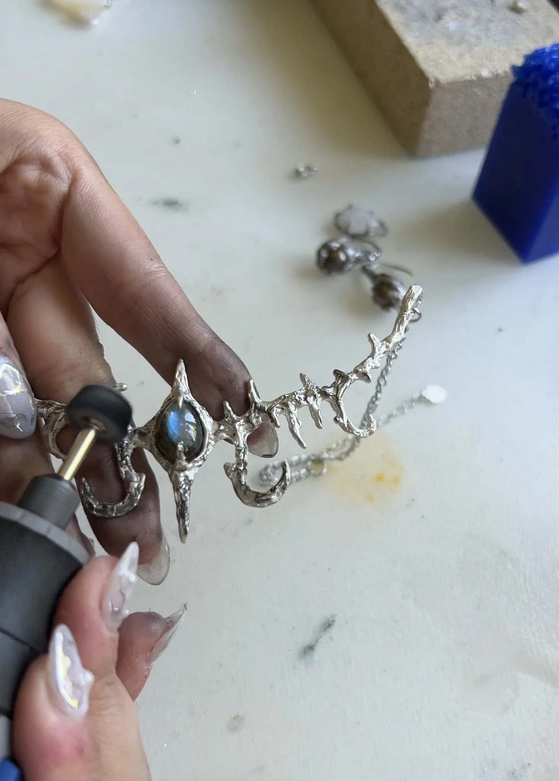 A person shaping a silver tiara or crown with intricate details, using a rotary tool on a work surface. The person’s fingers are dirty and their nails have a decorative design.