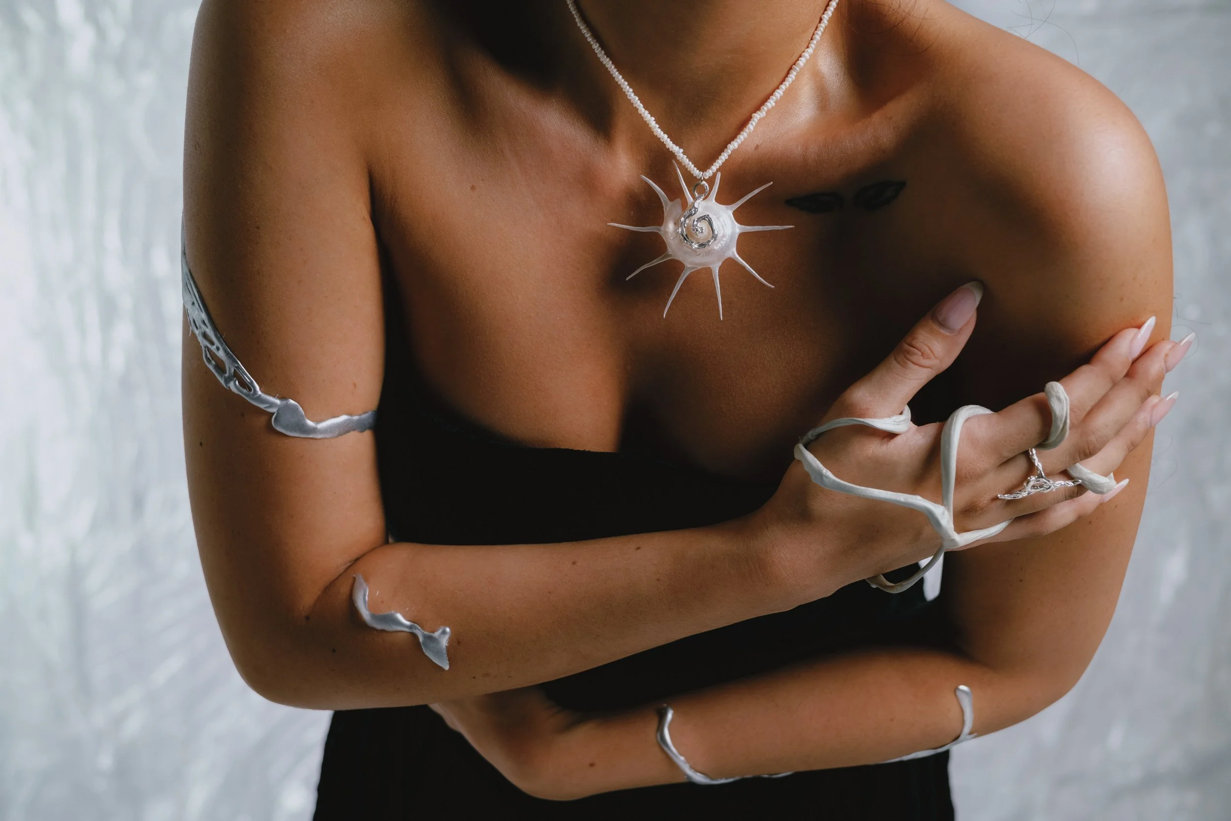 A woman wearing a silver sea star-shaped pendant, and matching jewelry on her arm, hand, and finger, crossing her arms against a textured grey background.