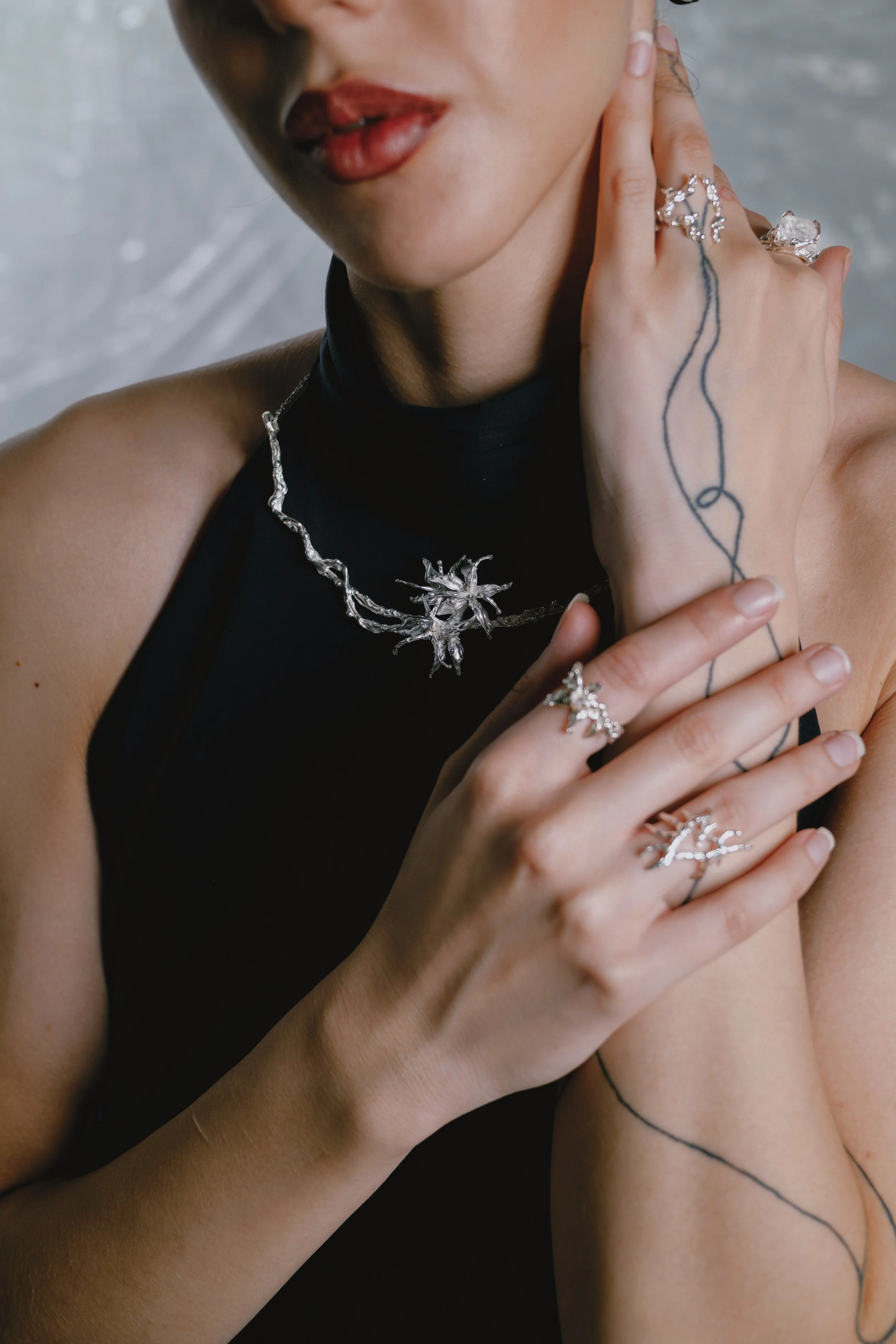 Close-up of a woman wearing silver jewelry, including a necklace and rings, with tattoos on her hand and arm, wearing a black sleeveless top.