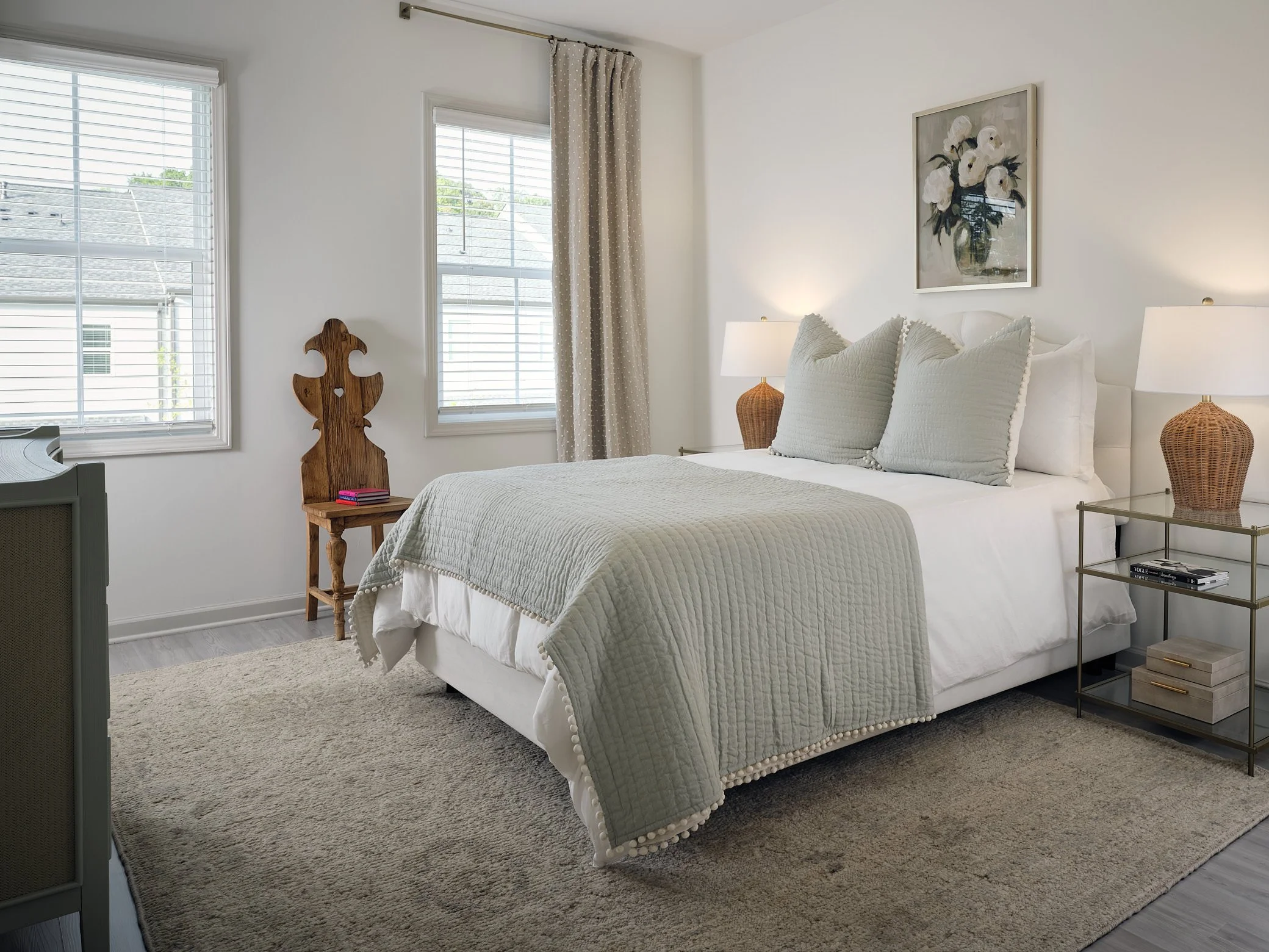 Bright bedroom with white walls, large bed with white linens and gray pillows, two side tables with lamps, two windows with beige curtains, a wooden chair, a decorative mirror, a framed floral artwork, and a beige rug.