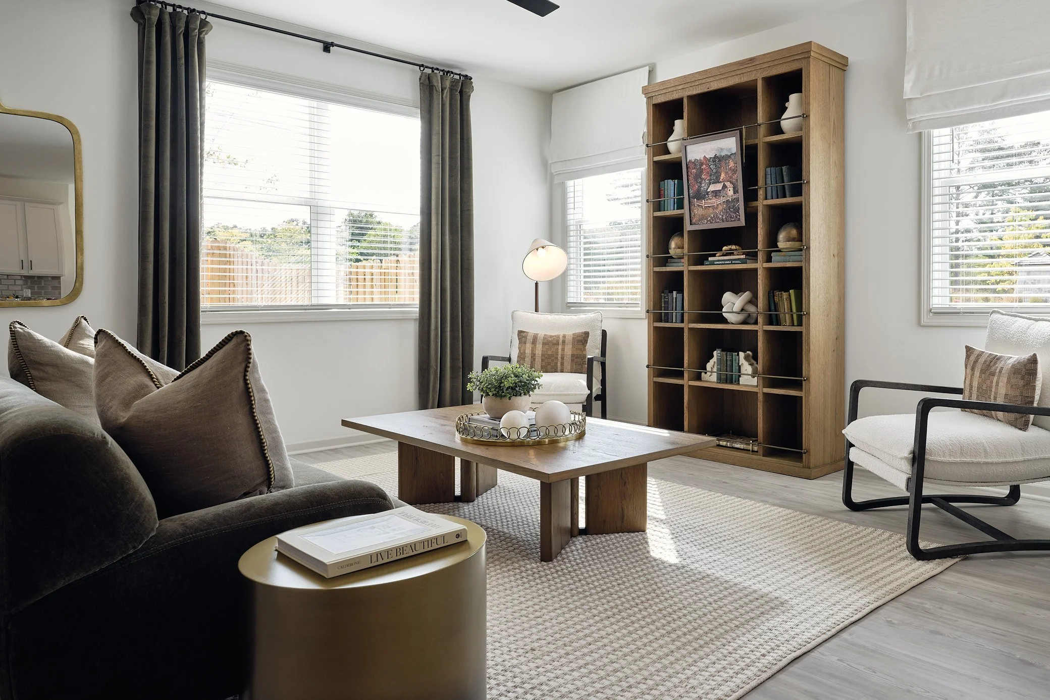 Open-concept living room with white walls, hardwood floors, a ceiling fan, and natural light from multiple windows, featuring a brown sofa, armchairs, coffee table, bookshelf.