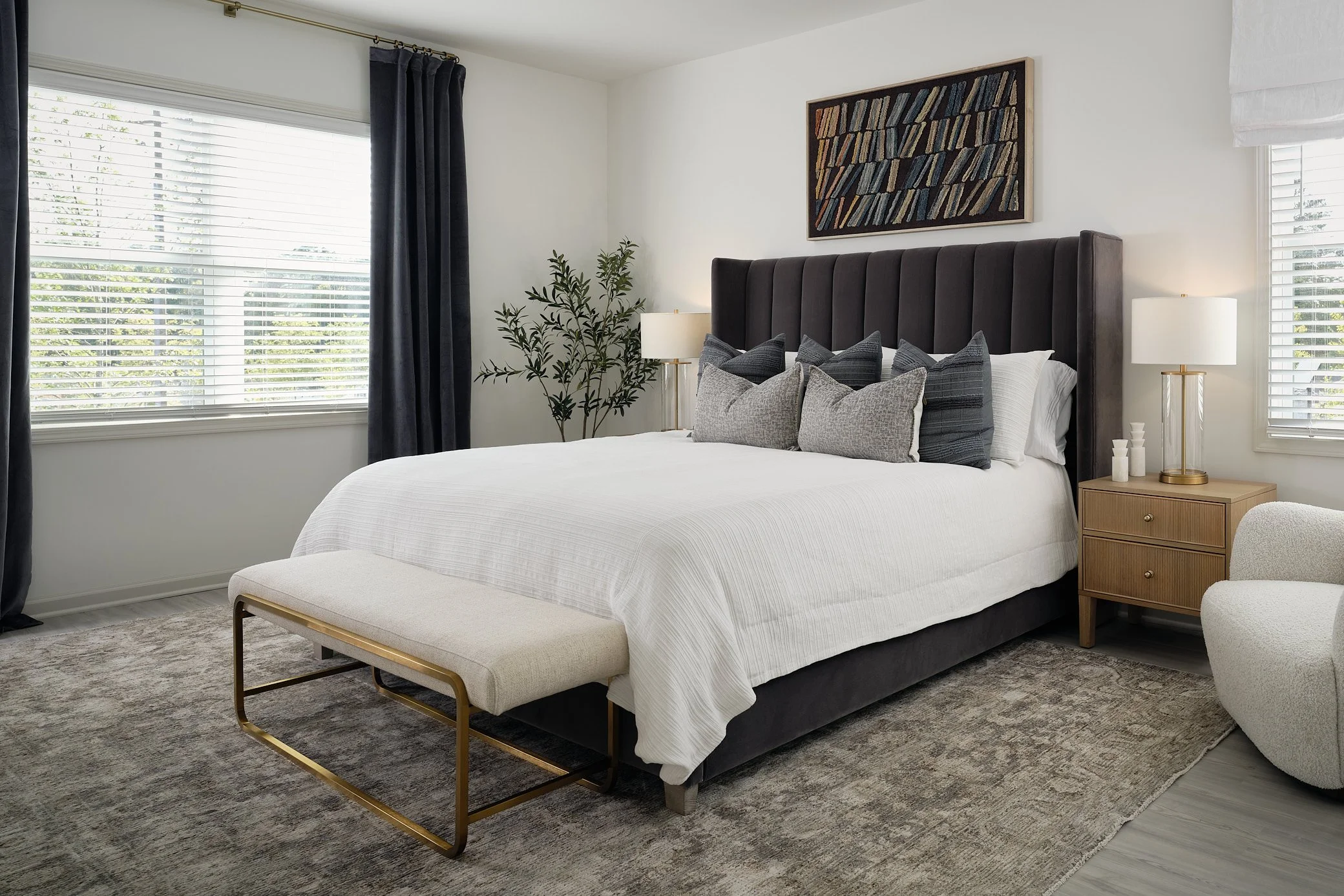 A modern bedroom with a large bed, gray upholstered headboard, multiple throw pillows, and a white bedspread. There are two nightstands with table lamps, a cream curved chair with a yellow pillow, a black ceiling fan, a window with blinds and dark cu