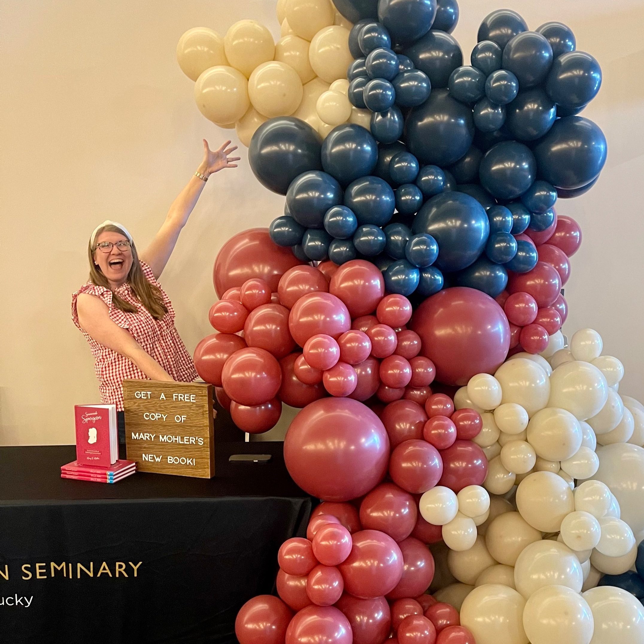 A woman in a red and white checkered dress is smiling and pointing at a multicolored balloon display at an event.