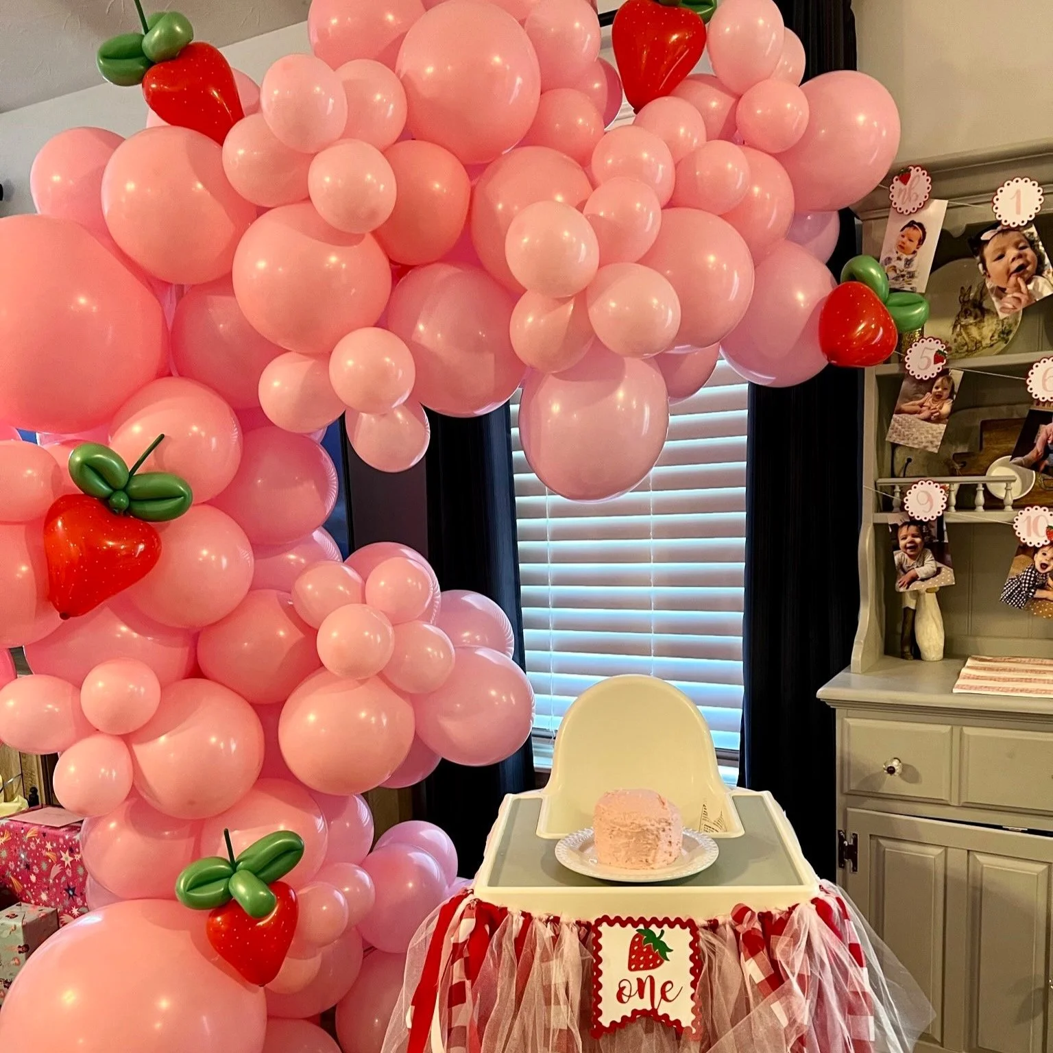 Decorative balloon arch with pink balloons and small strawberry-shaped balloons at a birthday celebration, featuring a high chair with a birthday cake and a sign that says 'one' with a strawberry illustration.