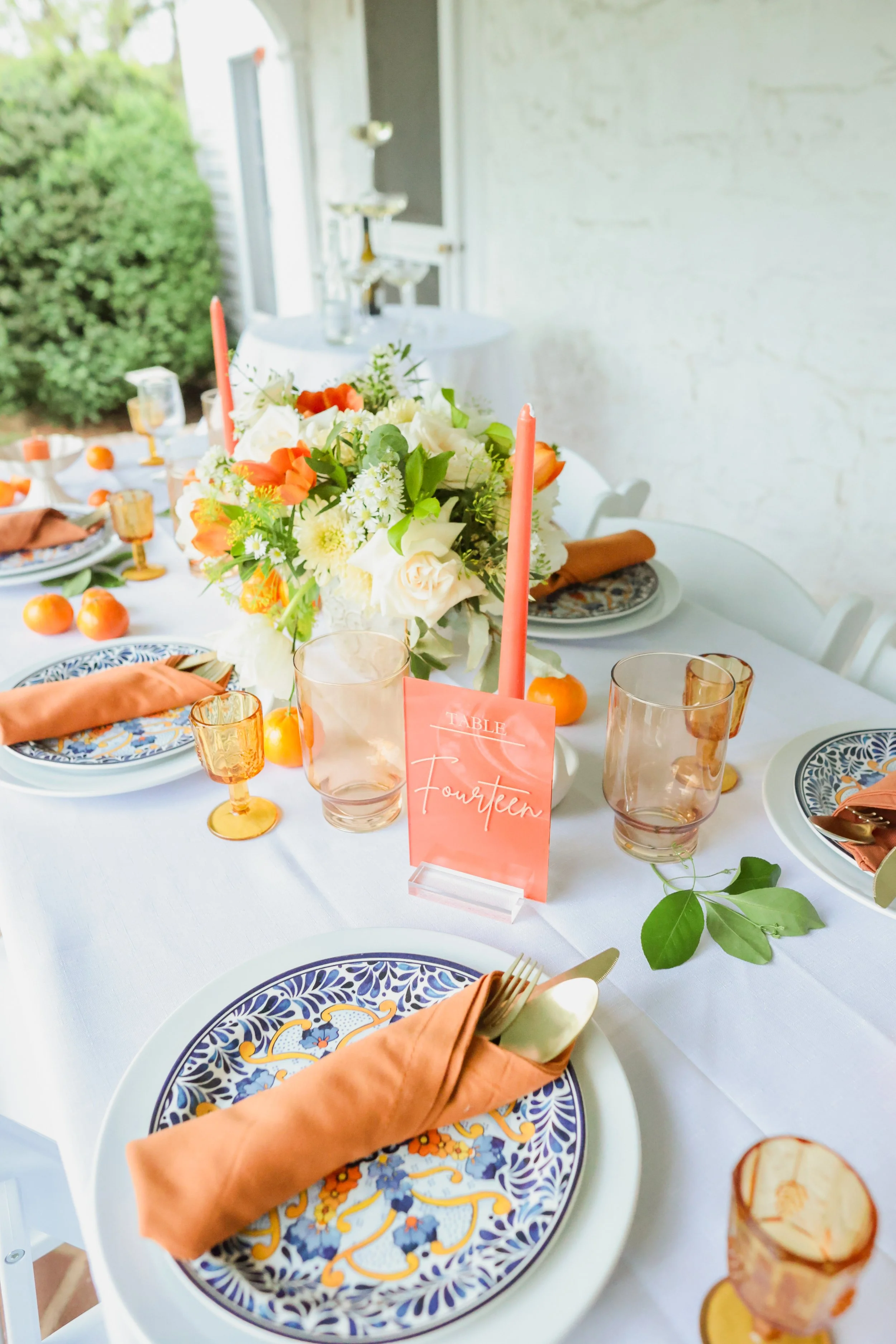 Set dining table decorated with floral centerpiece, coral candles, peach napkins, patterned plates, and glassware, with a table card labeled "Fourteen."