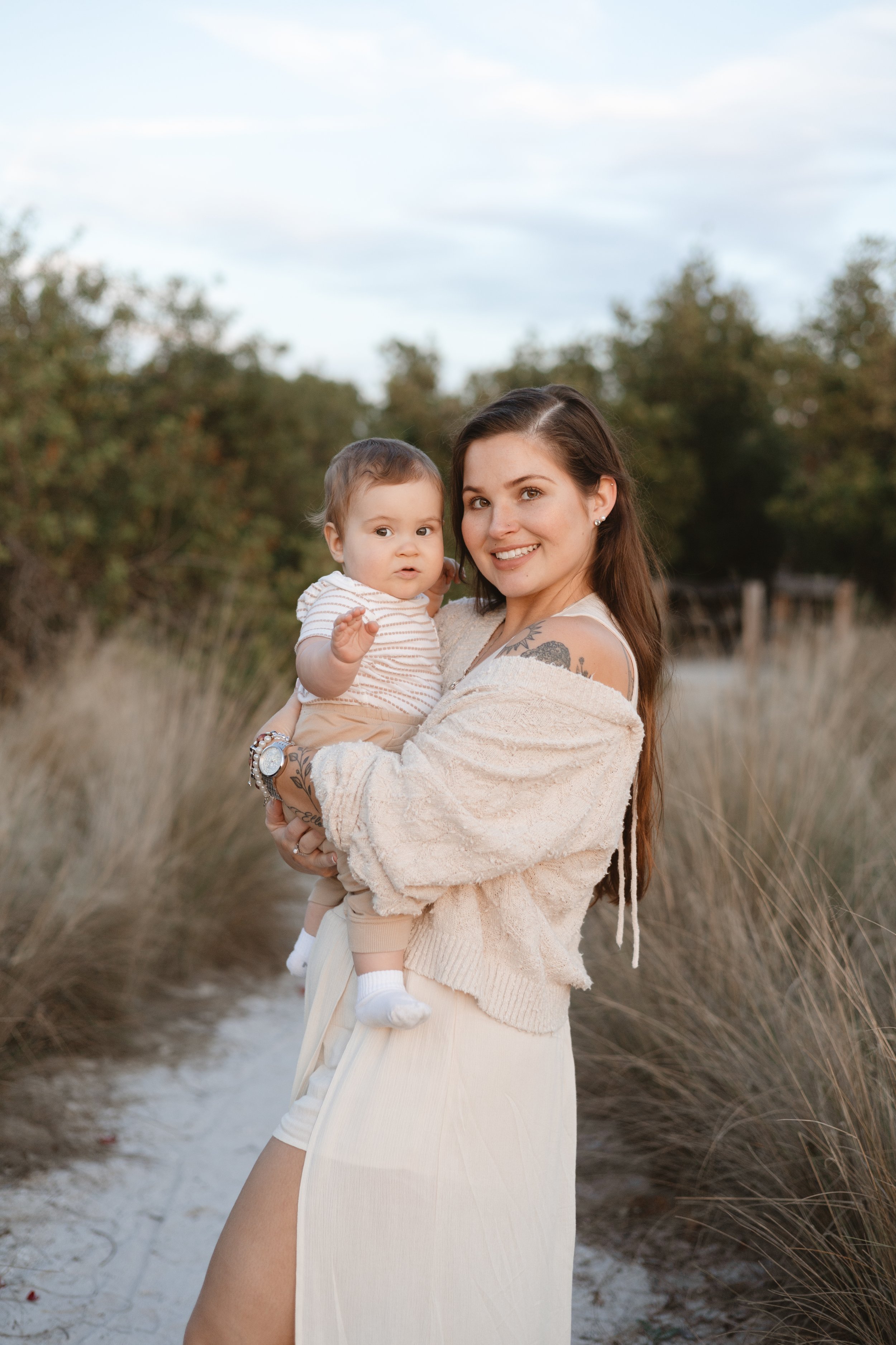 A smiling young woman with long brown hair holding a small child near a sandy pathway surrounded by tall grass and trees, under a partly cloudy sky.