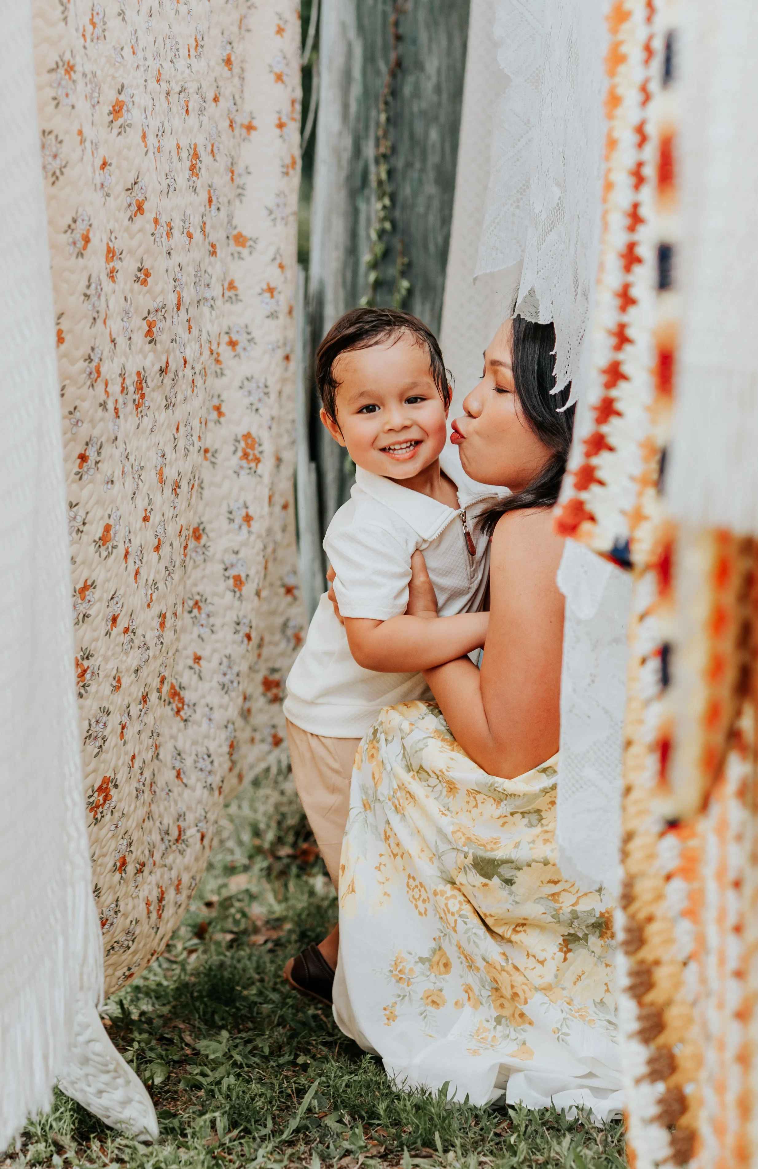 Mother kissing her smiling toddler while sitting between vintage quilts during a warm outdoor family photo session in Tampa, Florida.