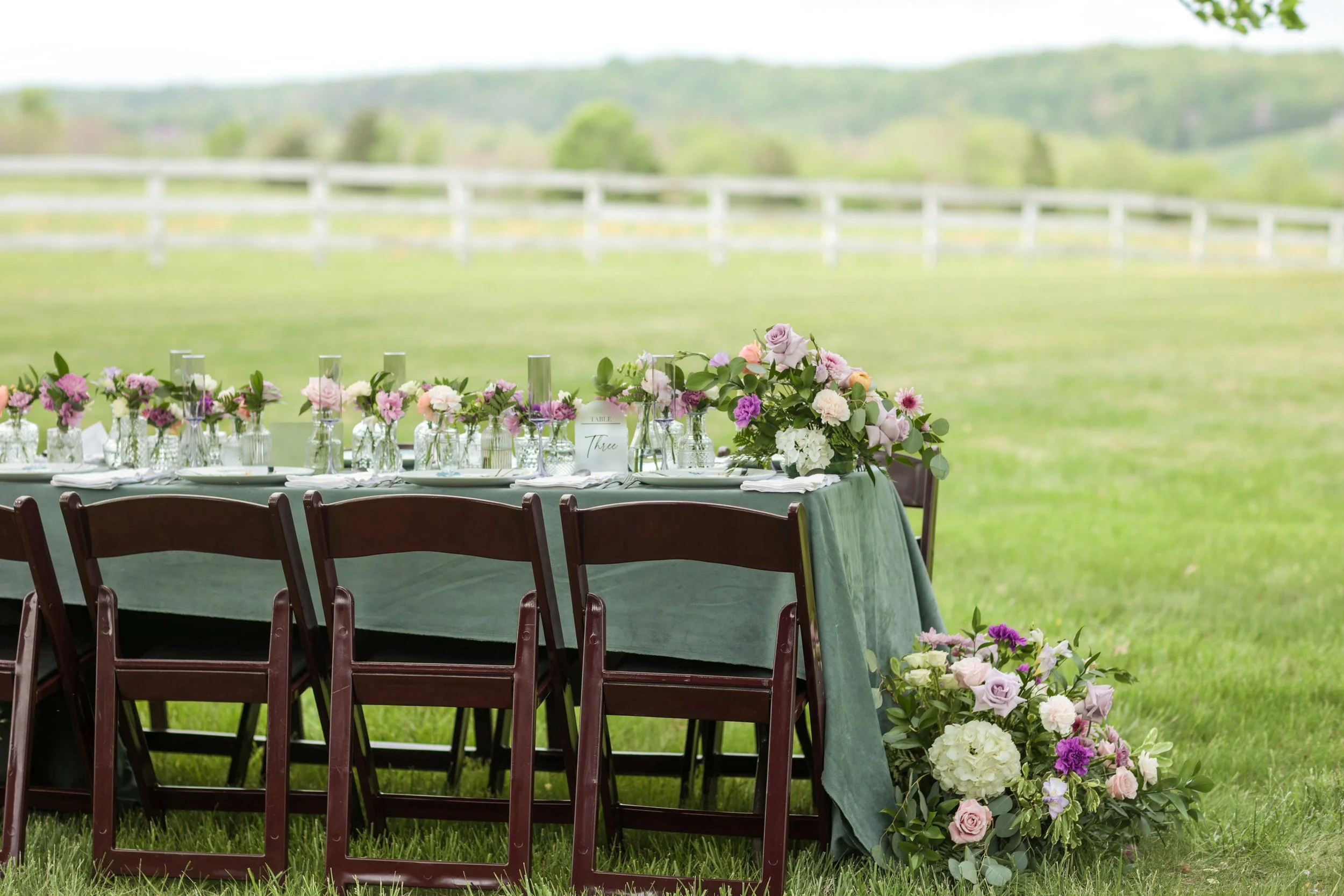 A table set for a celebration outdoors, decorated with pink, purple, and white flowers, and green tablecloths, on a grassy lawn with a white fence and rolling hills in the background.