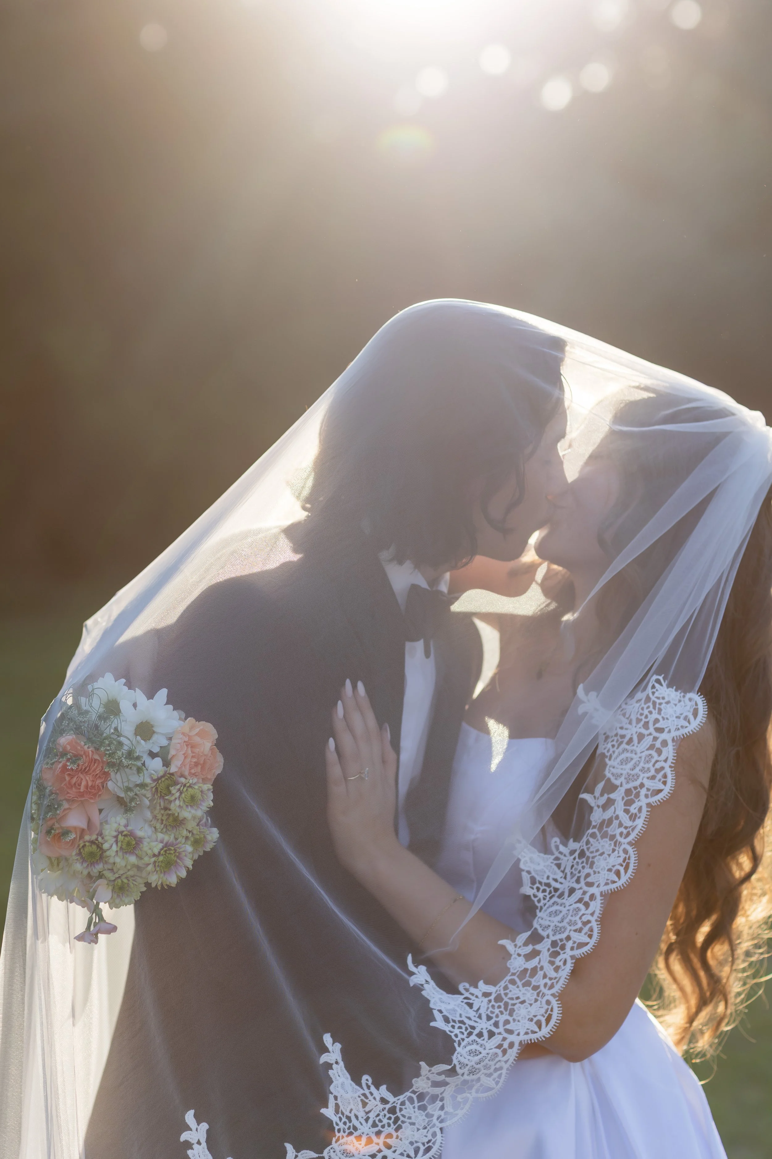 Backlit wedding portrait of a bride and groom sharing a kiss beneath a lace-trimmed veil during golden hour. Romantic and dreamy natural light photo with soft florals, perfect for timeless wedding photography inspiration.