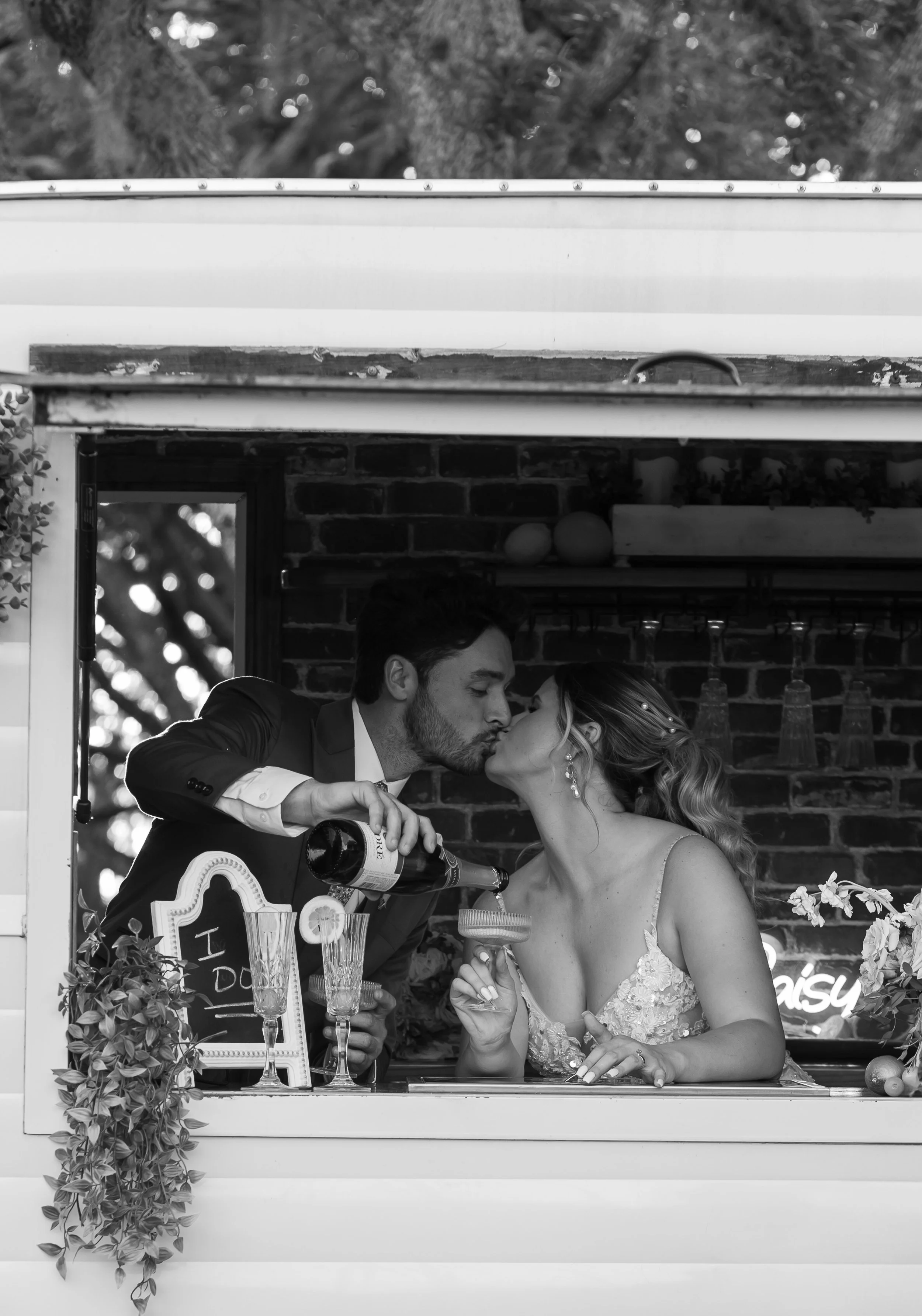 Black and white wedding photo of bride and groom sharing a romantic kiss inside a mobile bar trailer. Vintage wedding vibes with rustic details and candid, documentary-style photography—perfect for modern yet timeless love stories.
