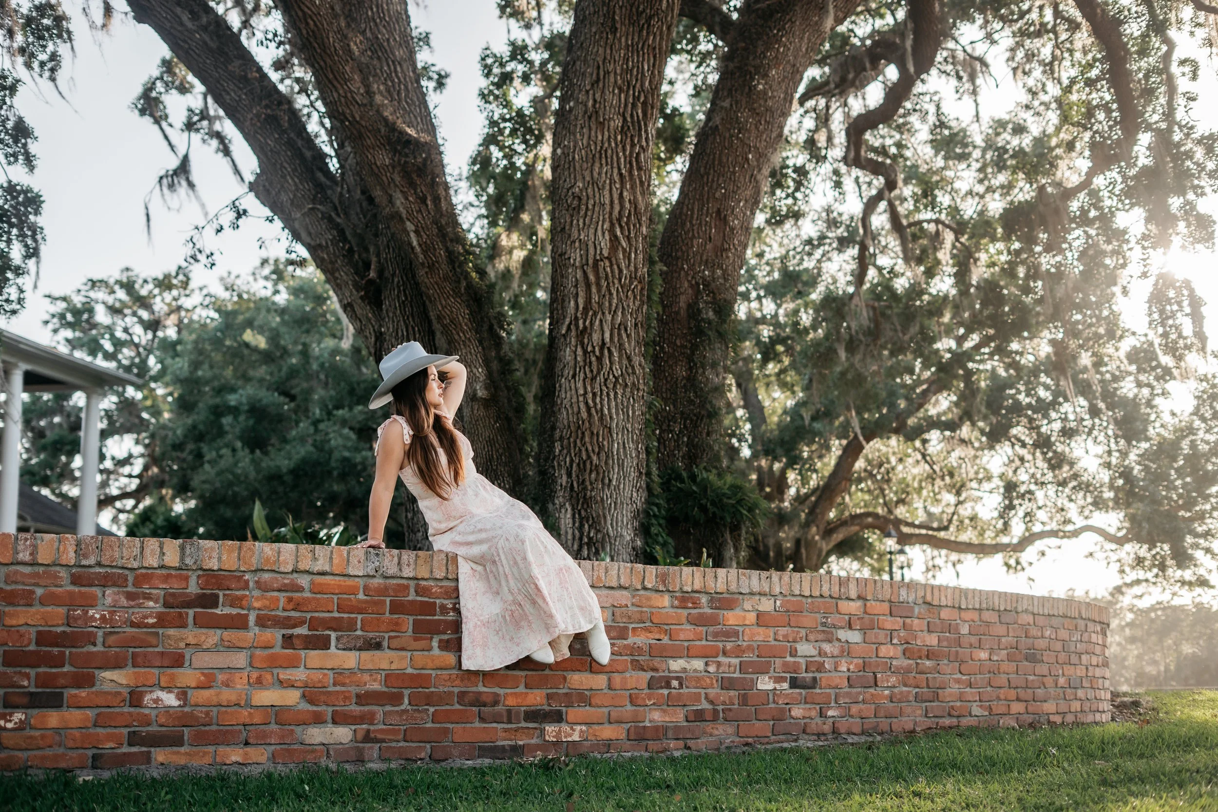 Woman in a floral dress and wide-brimmed hat sitting on a brick ledge beside a large tree in a park during sunset.