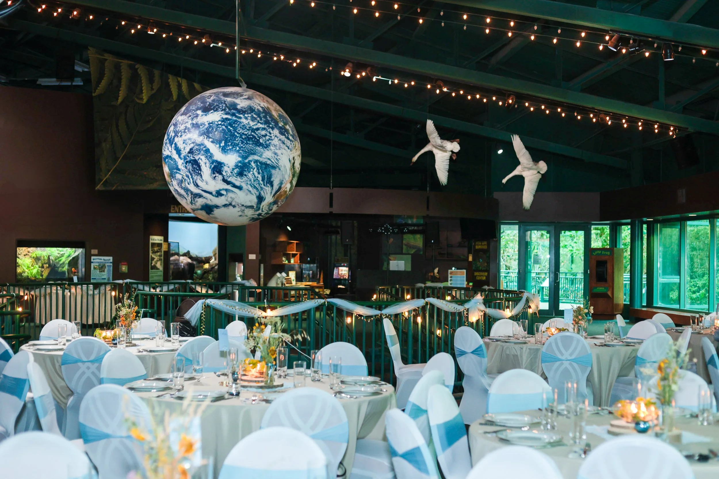 Event space decorated with white and blue chairs, tables with floral centerpieces, and a large hanging globe of Earth with string lights overhead. Decorative white birds hang from the ceiling, and there is a large window showing greenery outside.