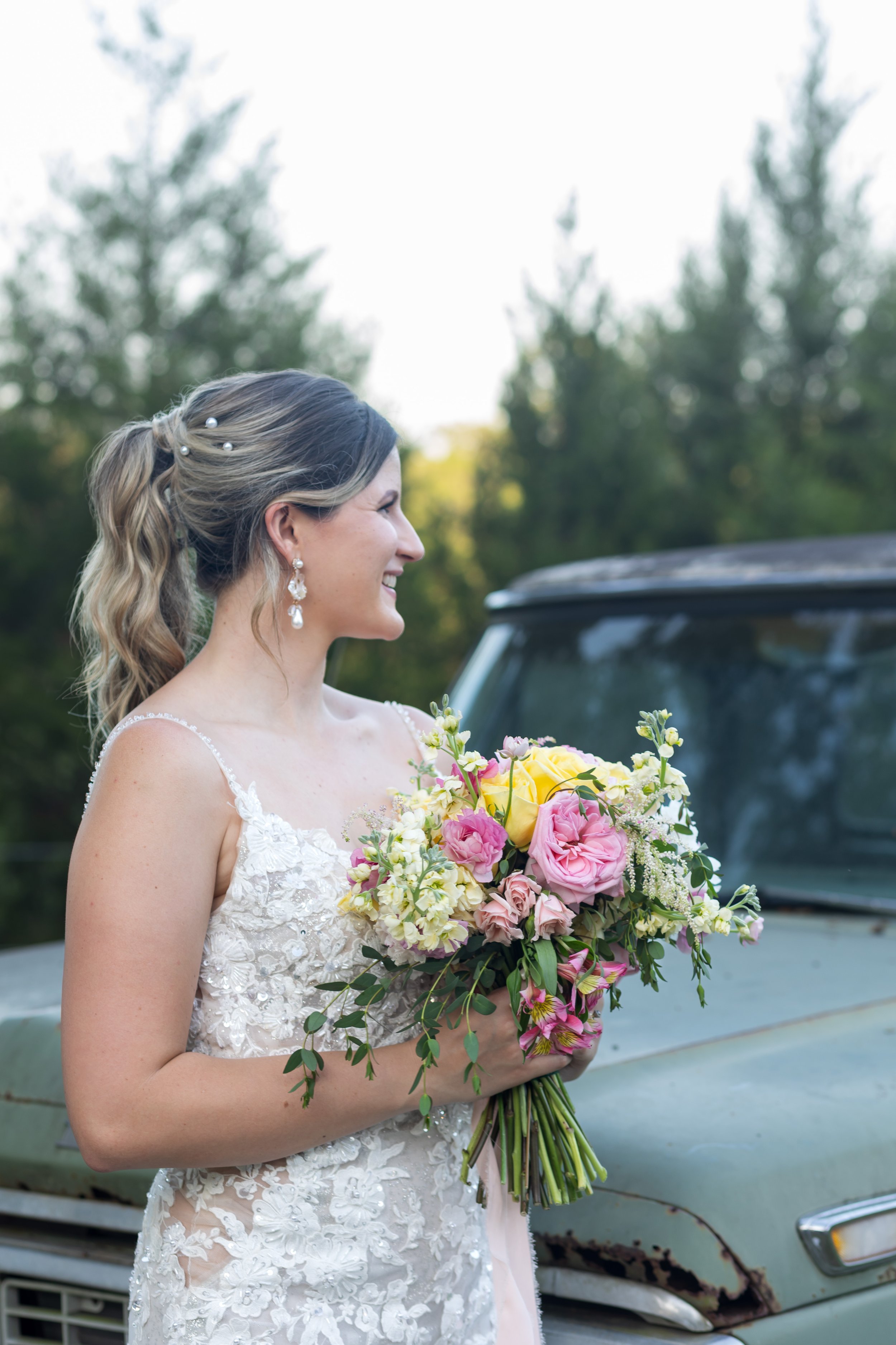 Outdoor bridal portrait of a bride in a lace wedding gown holding a colorful bouquet with pink, yellow, and white flowers. Captured in front of a vintage green truck with a backdrop of pine trees—perfect for rustic or country wedding inspiration.