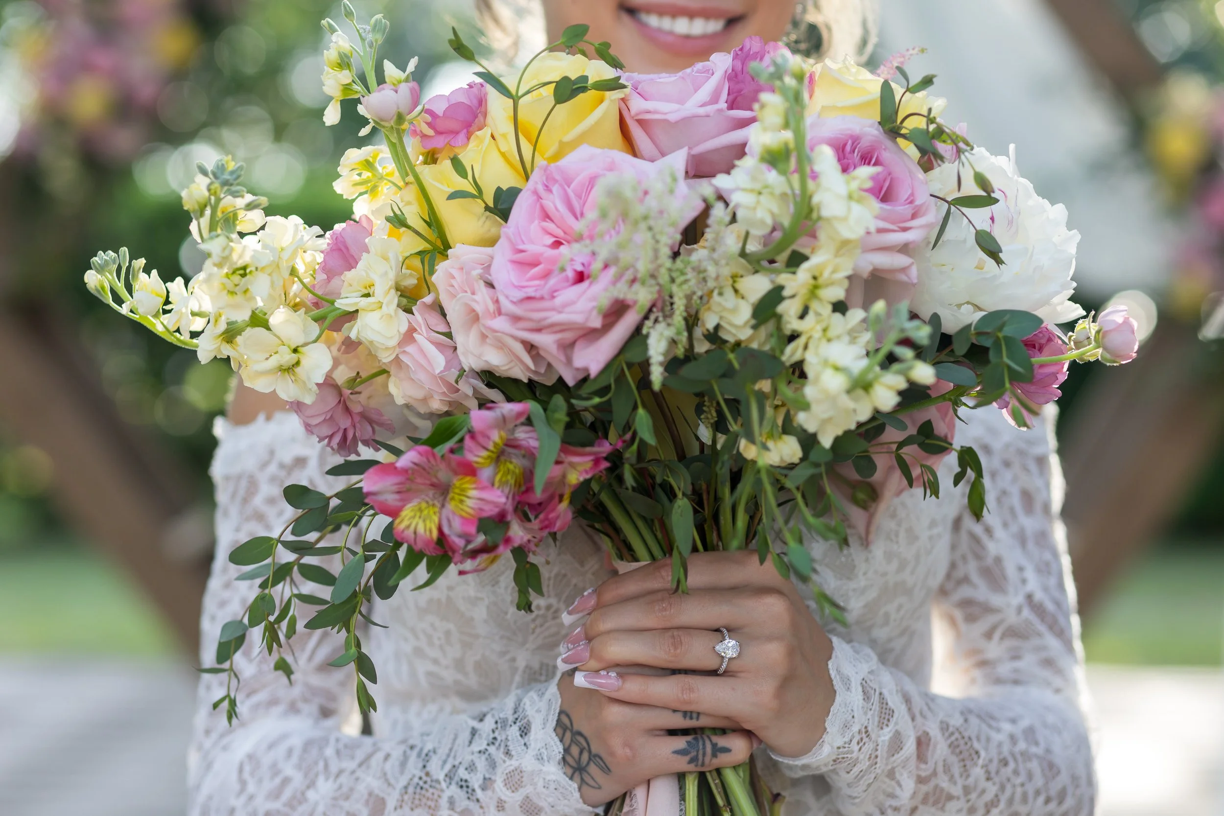 Bride holding vibrant spring wedding bouquet with pink peonies, yellow roses, and snapdragons. Lace wedding dress and sparkling engagement ring visible in close-up floral portrait—perfect for romantic garden wedding inspiration.