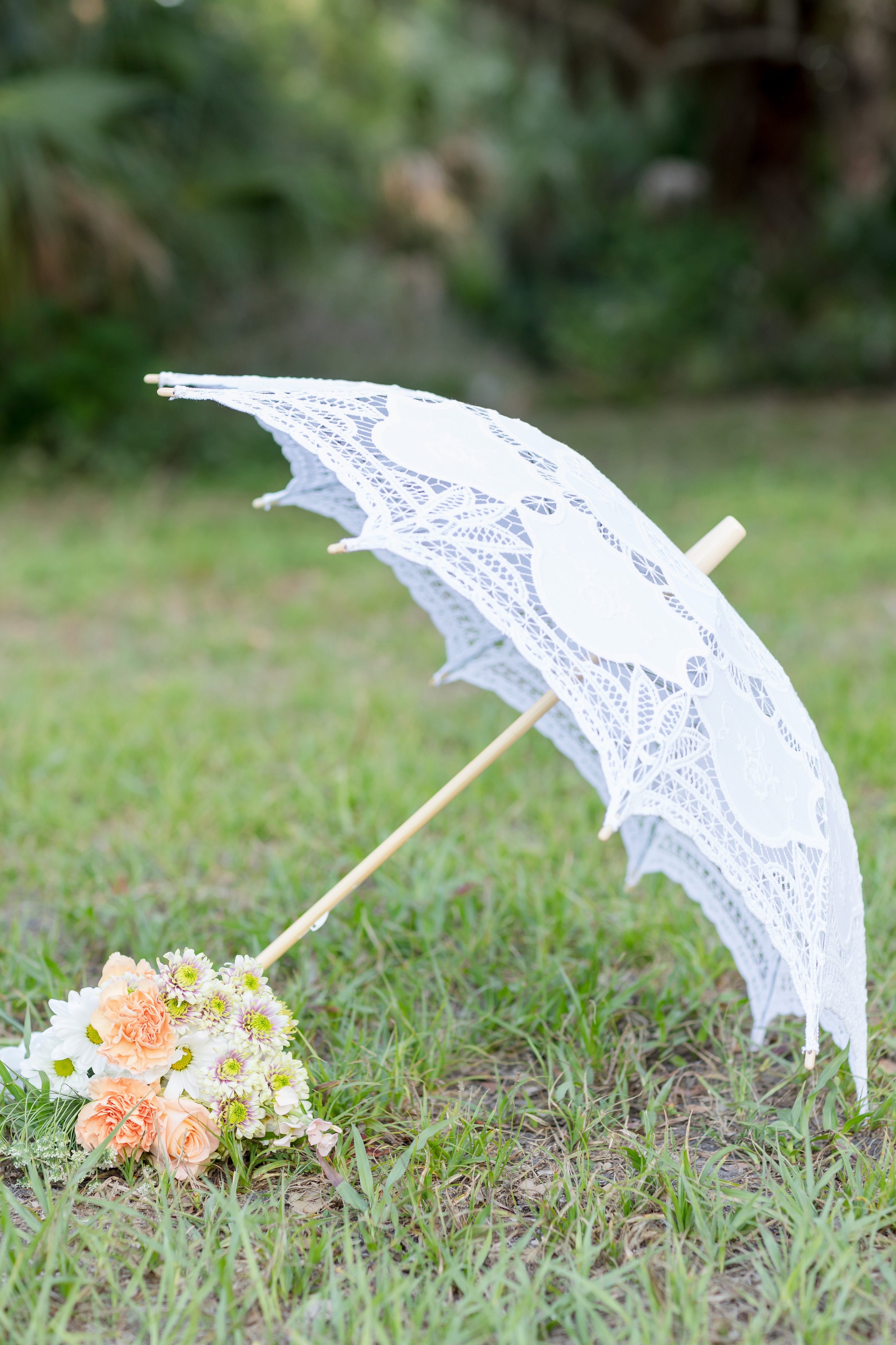 Romantic outdoor wedding detail featuring a white lace parasol and pastel bridal bouquet resting in the grass. Perfect for vintage garden wedding inspiration, Southern charm themes, or spring bridal styling ideas.